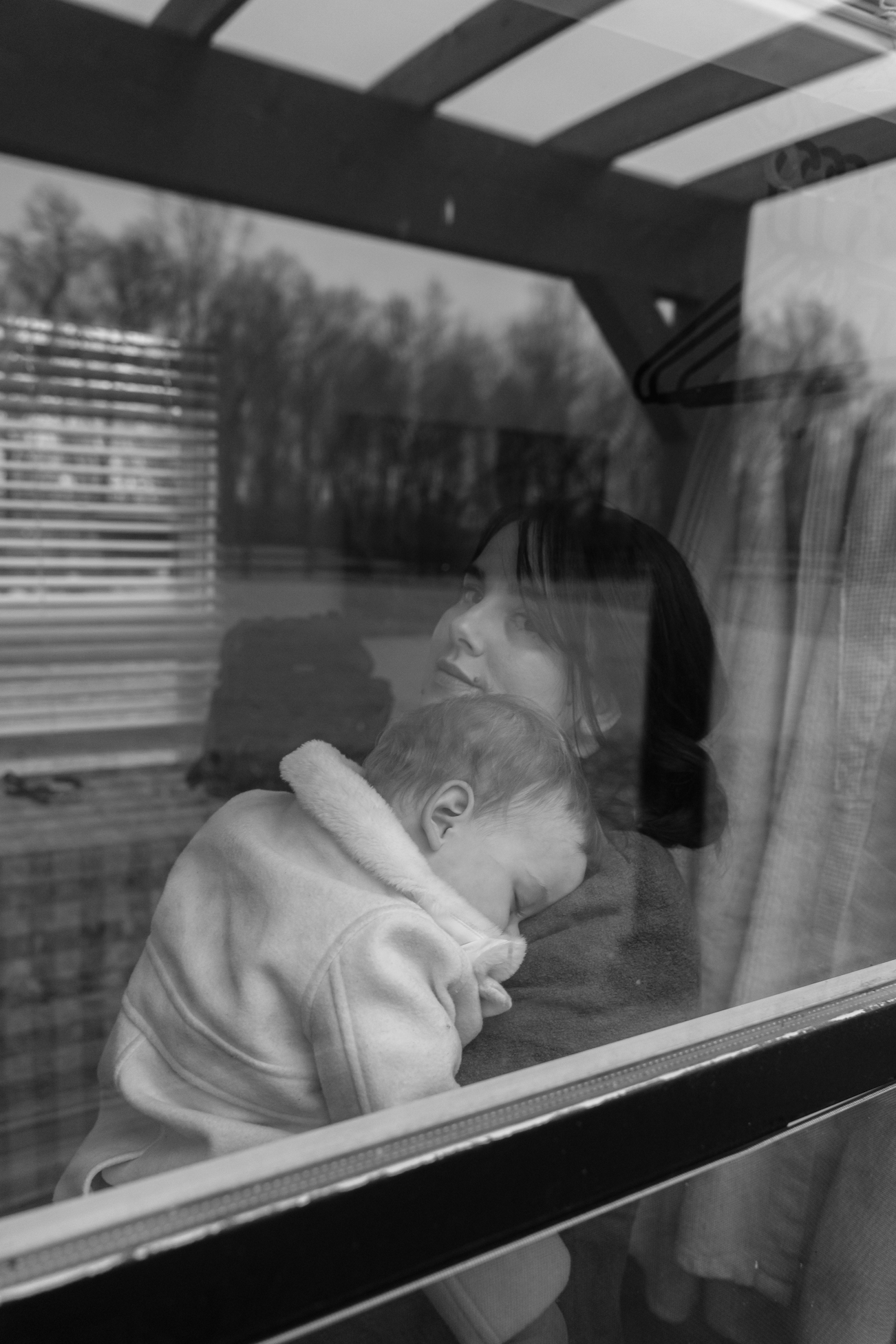 A woman and a young boy are seen through a window, with the boy resting his head on her shoulder. The scene appears to be outside, possibly in a backyard or park, with trees and horizontal fencing visible in the background. The photograph is in black