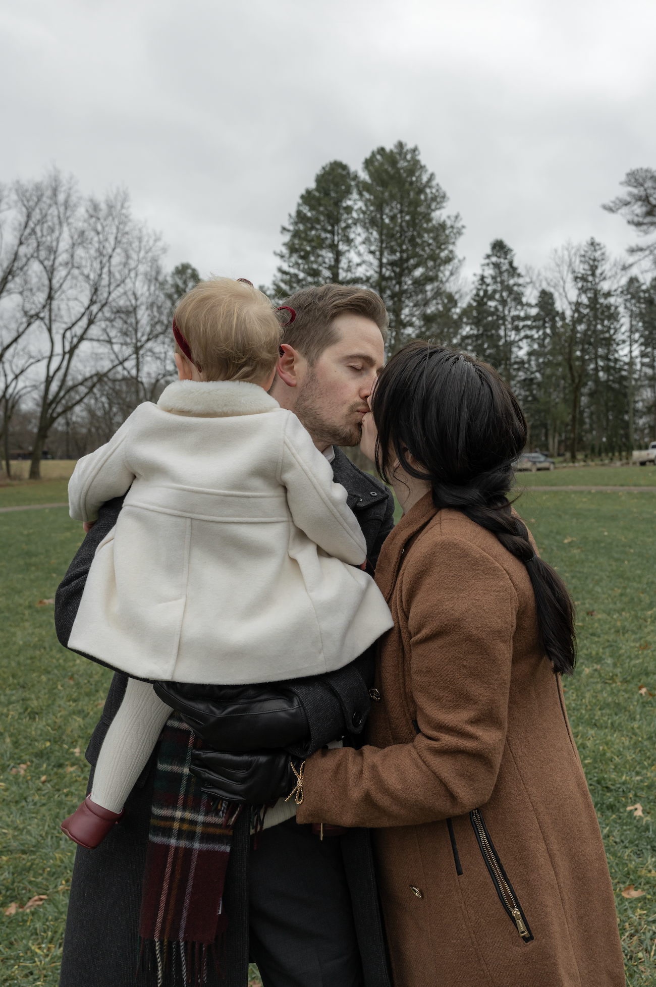 A couple sharing a kiss outdoors while holding a small child in a park with trees and cloudy sky in the background.