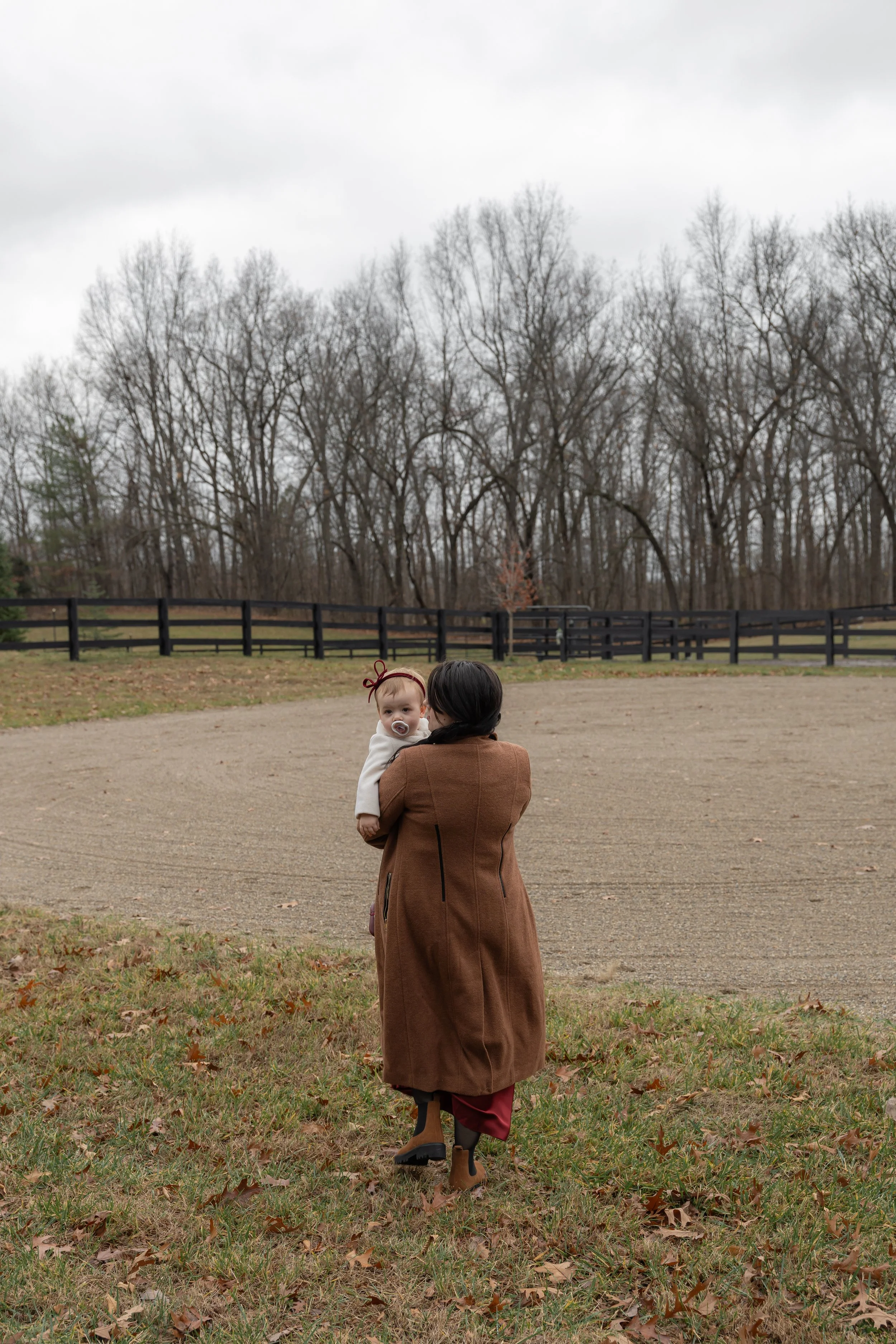 A woman holding a baby outdoors in a field with a black wooden fence and trees in the background on a cloudy day.