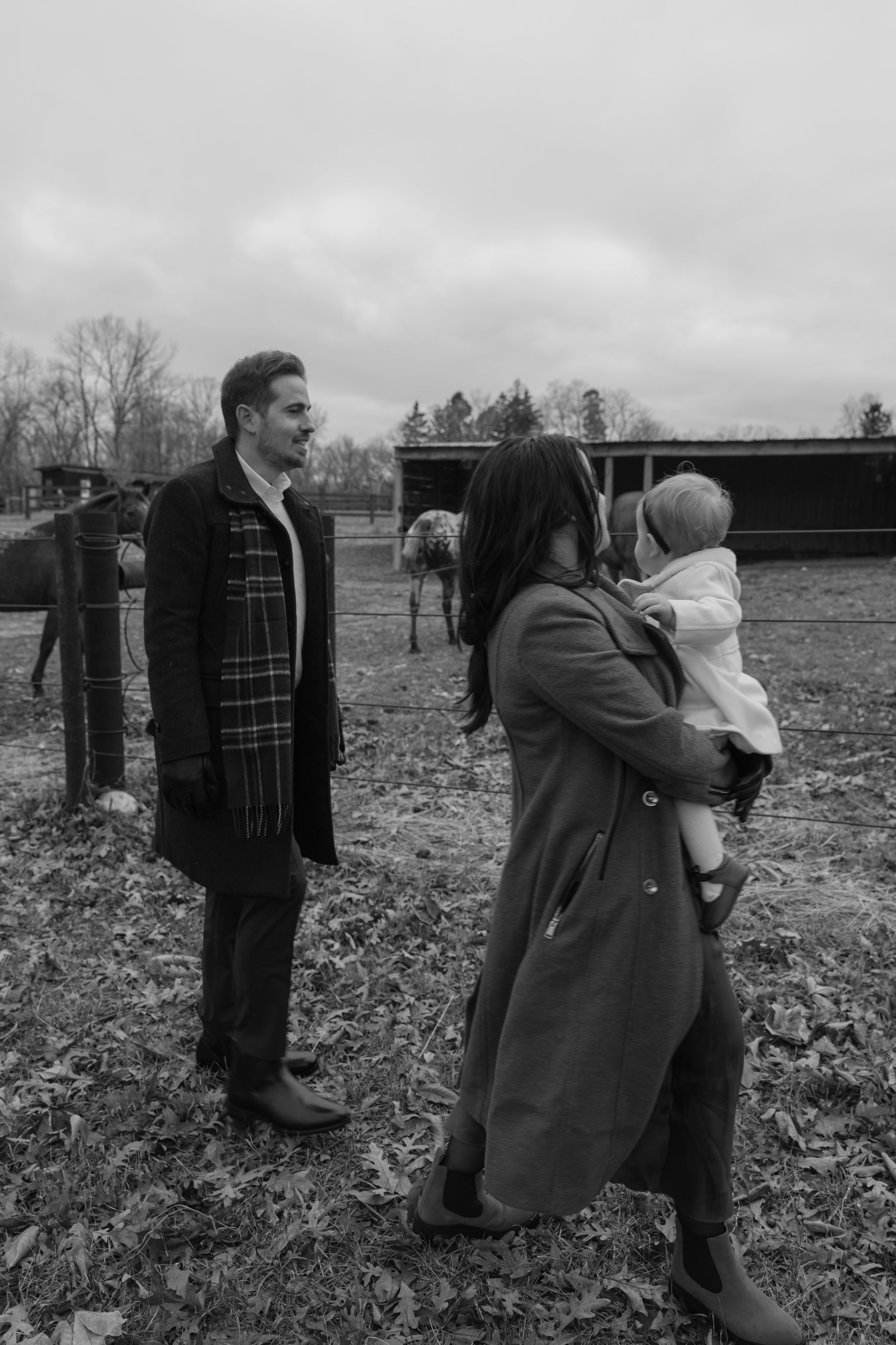 A woman holding a child in front of a fence with horses and a barn in the background on a cloudy day