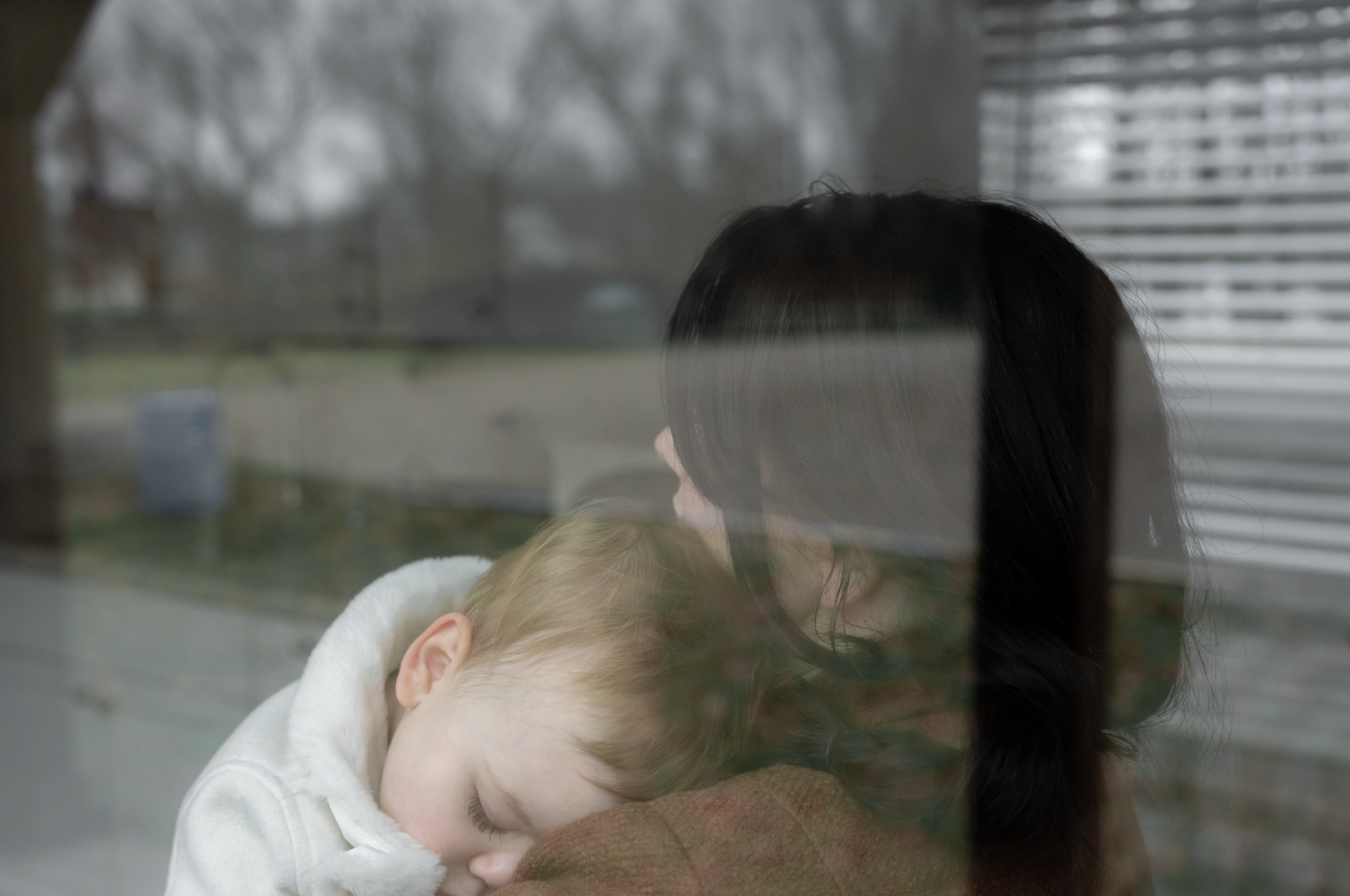 A young child with blonde hair sleeping on an adult's shoulder, seen through a window with a blurred outside view.