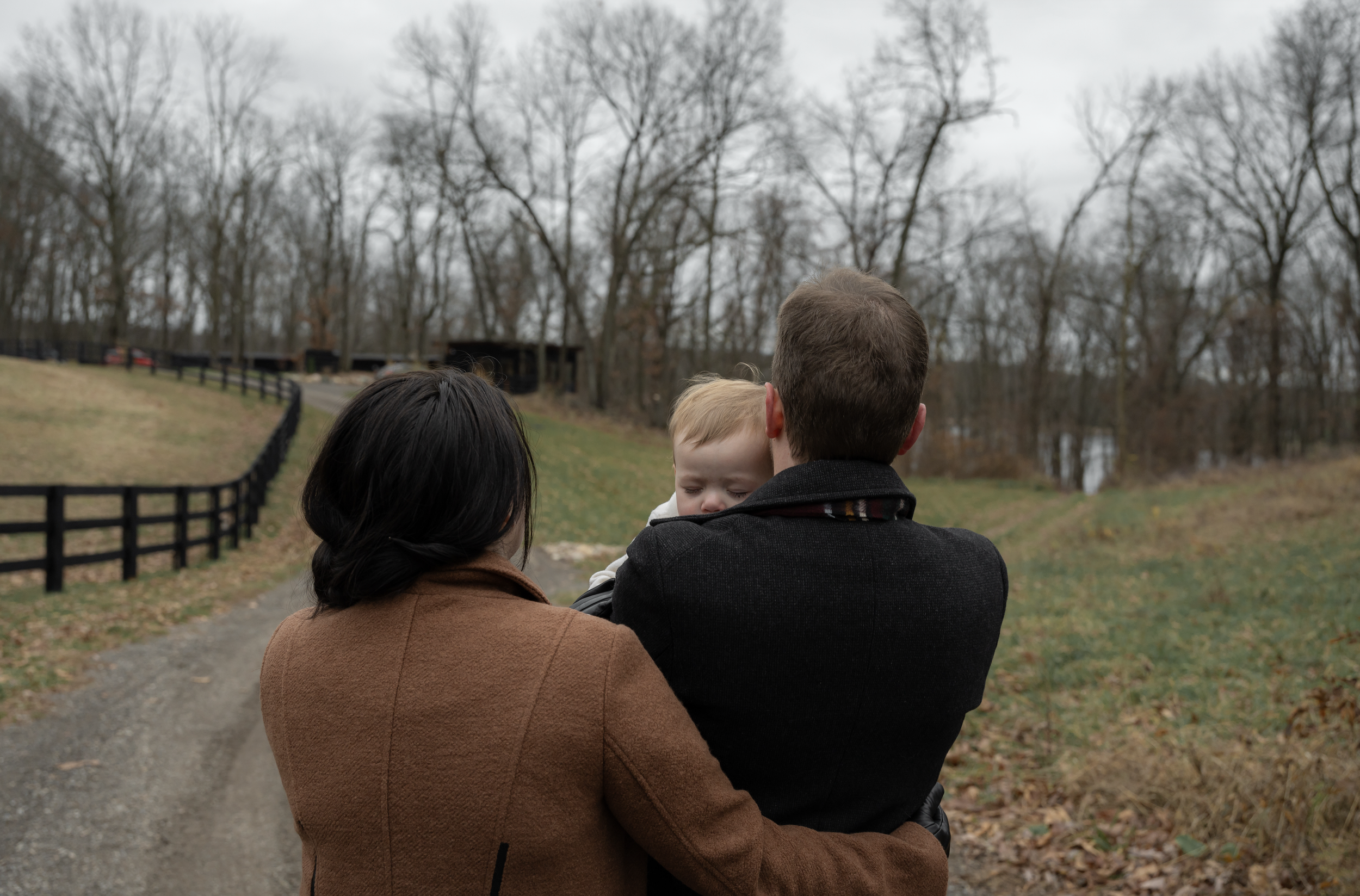 A woman, man, and baby are outdoors during fall or winter with leafless trees and cloudy sky in the background. The woman has dark hair and is wearing a brown coat. The man has short hair and is holding the baby, who has blonde hair and is looking do