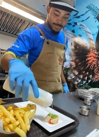 A man wearing a Lionfish Caribbean apron and a cap, holding a lionfish, standing in a kitchen with a fish-themed mural in the background.