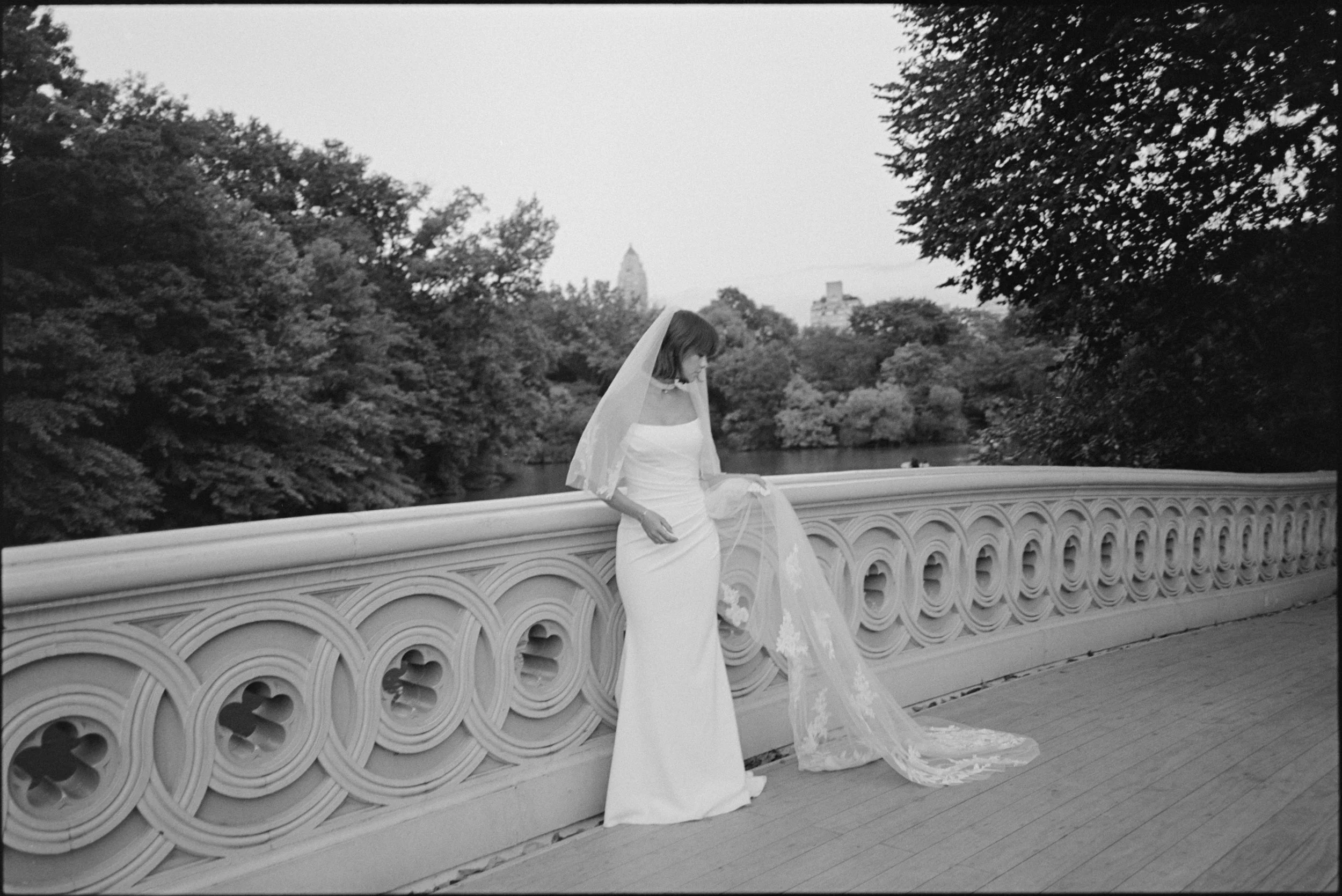 Black and white photo of a woman in a wedding dress with a long train and veil, standing on a bridge with an ornate railing, overlooking a park with trees and a silhouette of a city skyline in the background.