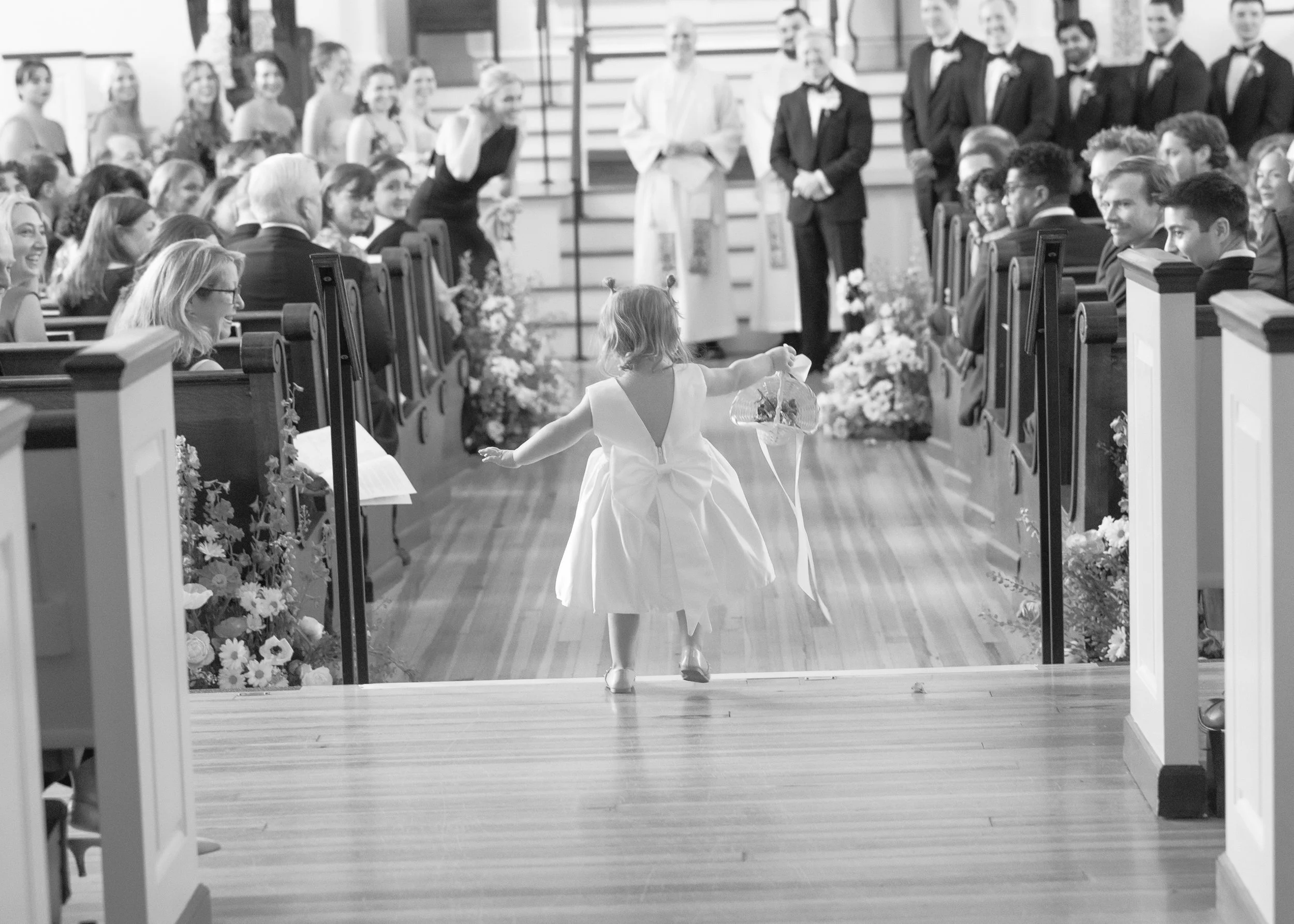 A young girl in a white dress walking down the aisle of a church during a wedding, holding a basket.