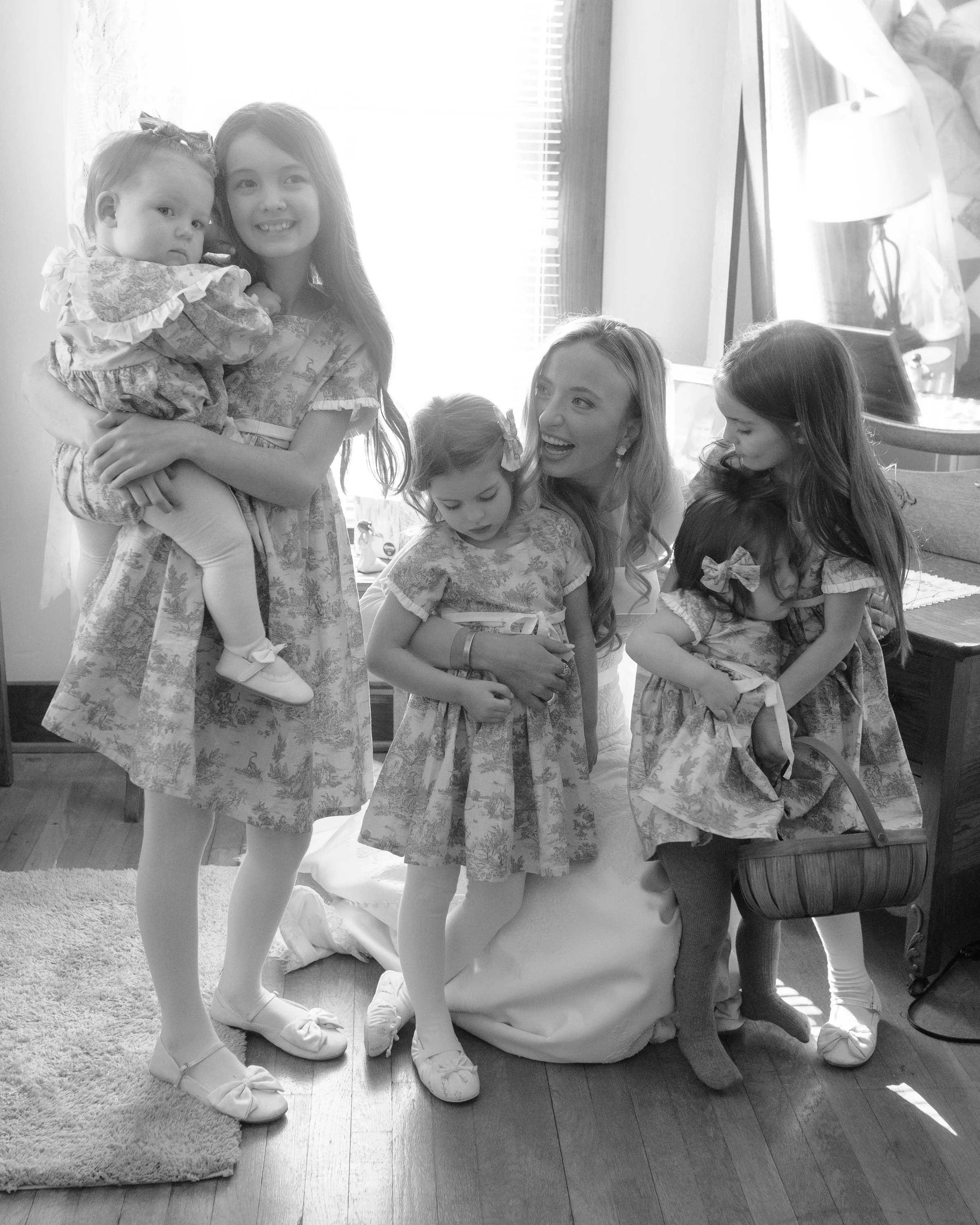 Group of young girls, four standing and three sitting, wearing floral dresses with bows in their hair inside a well-lit room with a window and a lamp.