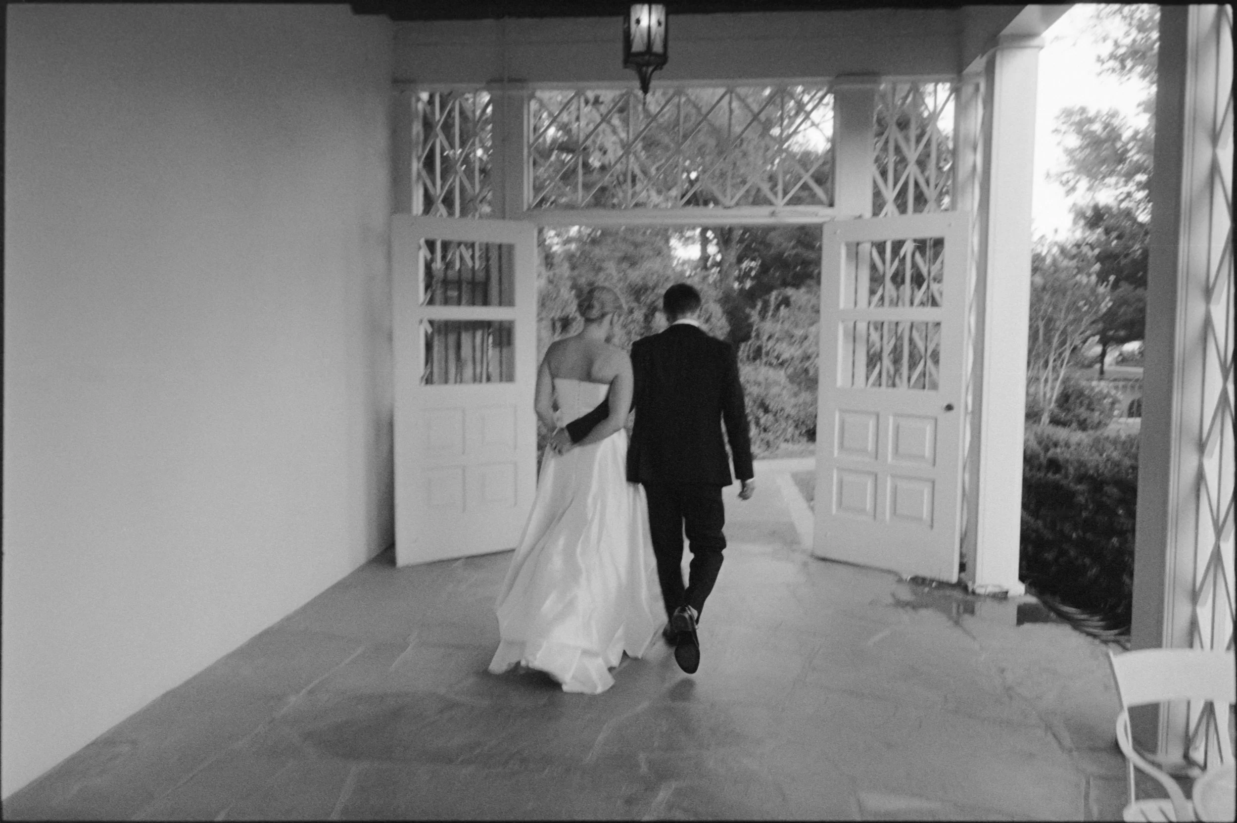 Black-and-white photo of a bride in a wedding gown and a groom in a suit walking out of a building, holding hands, with open double doors leading outside.
