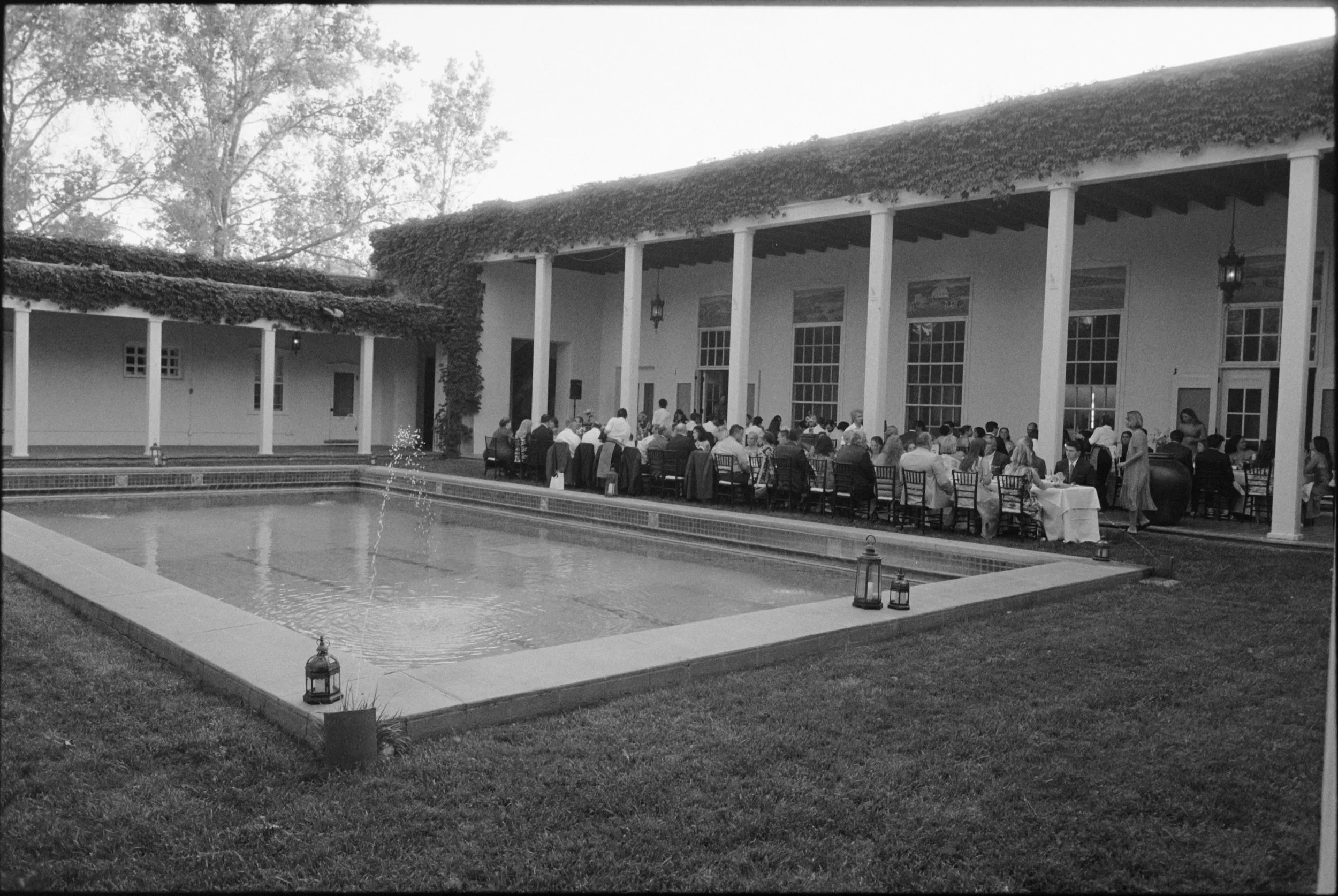 A large outdoor dining area beside a building with tall white columns, large windows, and a roof covered in vines. Guests are seated at tables under the columns, and lanterns are placed along the edge of a fountain in the foreground.