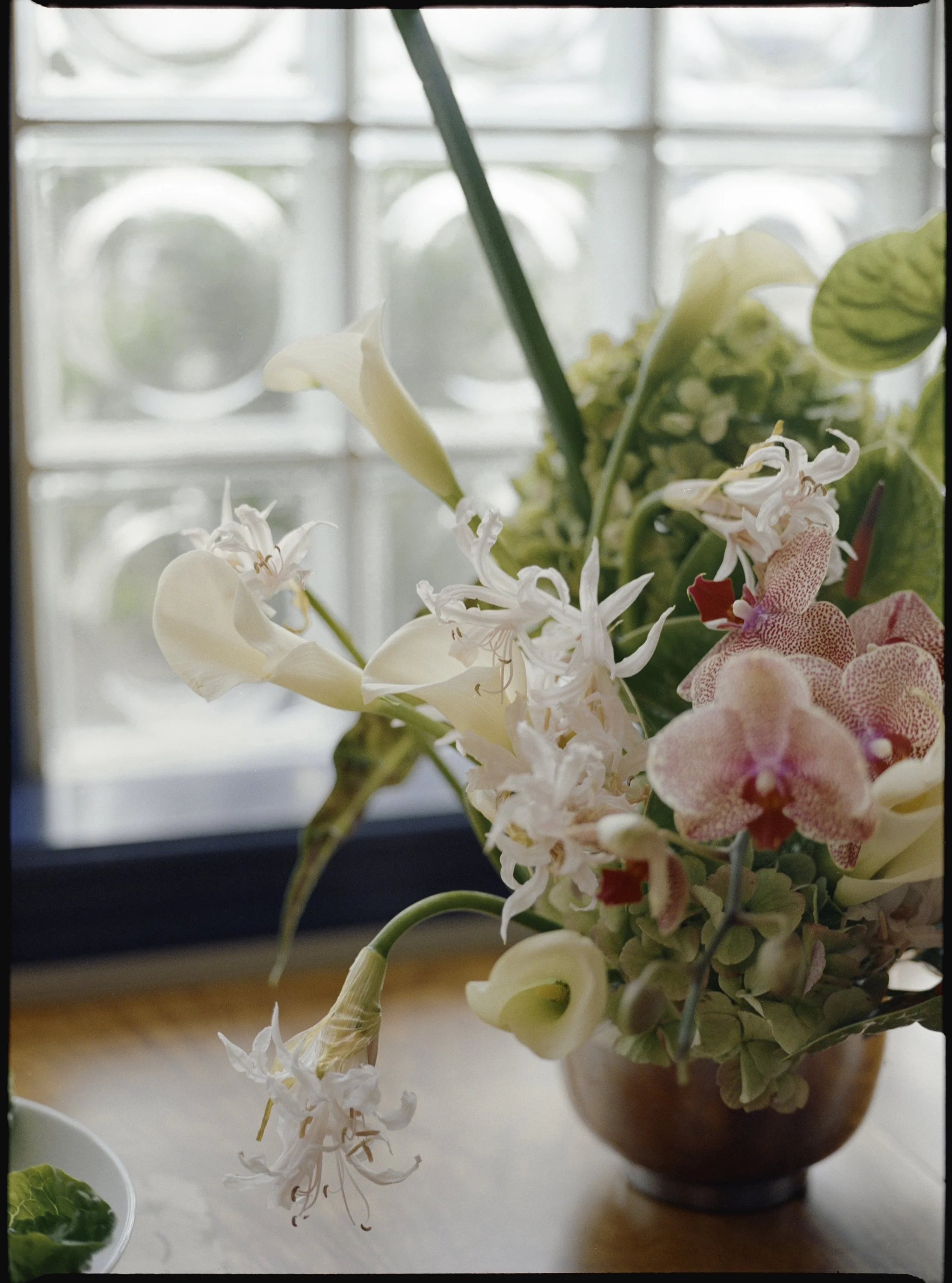A floral arrangement in a paper brown vase on a wooden surface near a window with frosted glass. The arrangement includes white calla lilies, pink and white orchids, and green leaves.