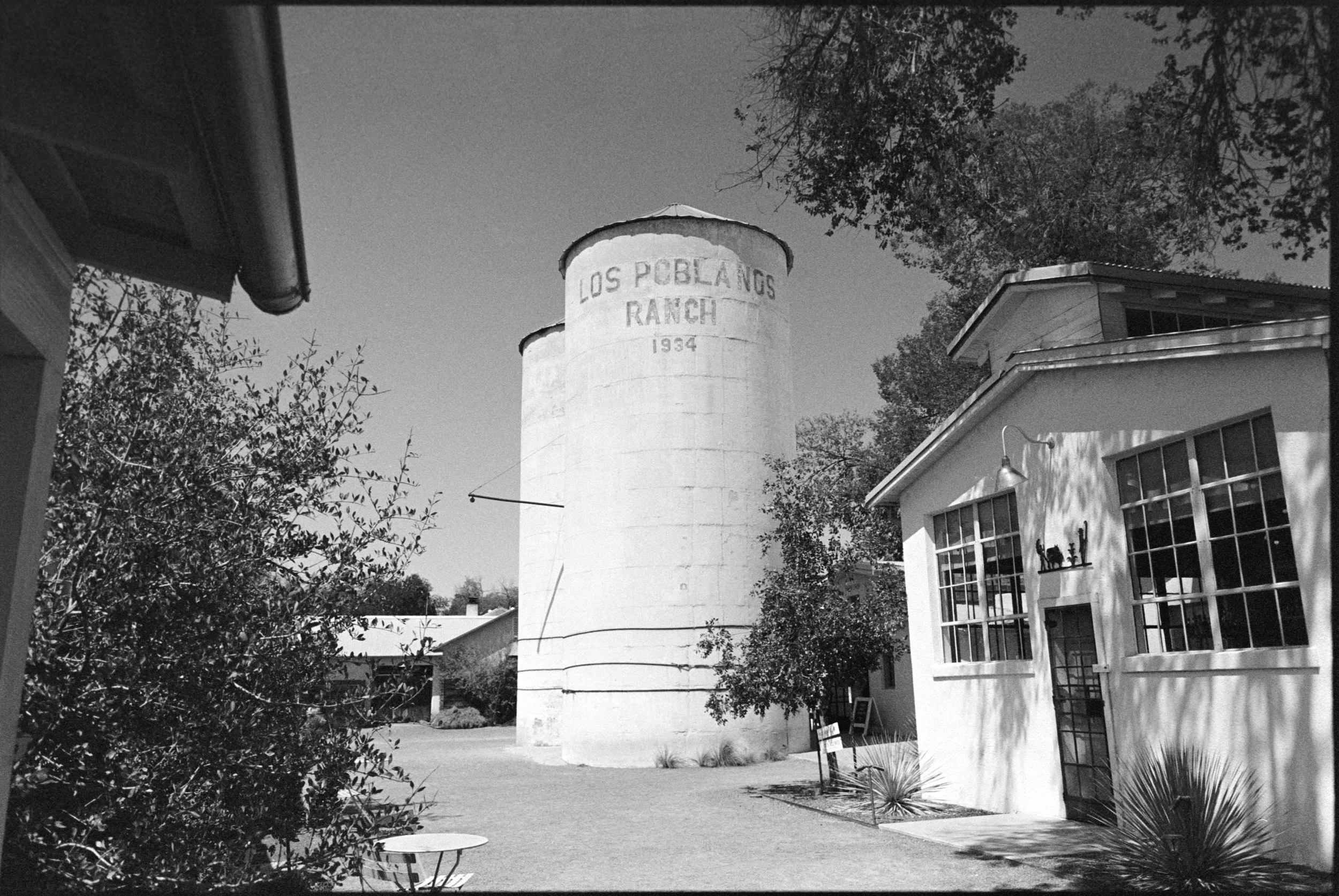 A black and white photo of a traditional water tower with the text "Los Pobladores Ranch 1934" written on it, surrounded by trees and modern buildings.