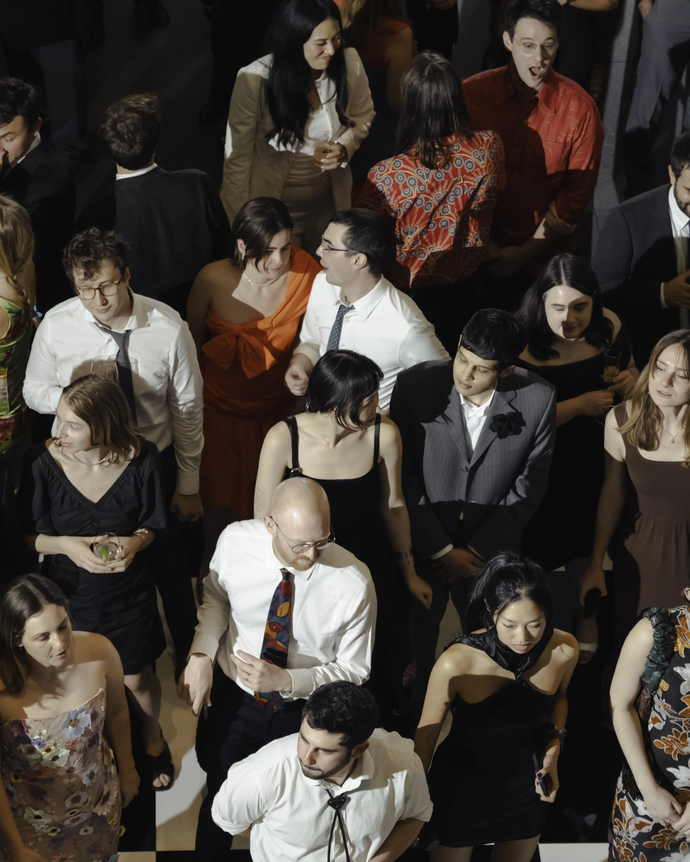 People gathered at a social event, engaging in conversations, dressed in formal and semi-formal attire, with some holding drinks. The setting appears to be an indoor venue.