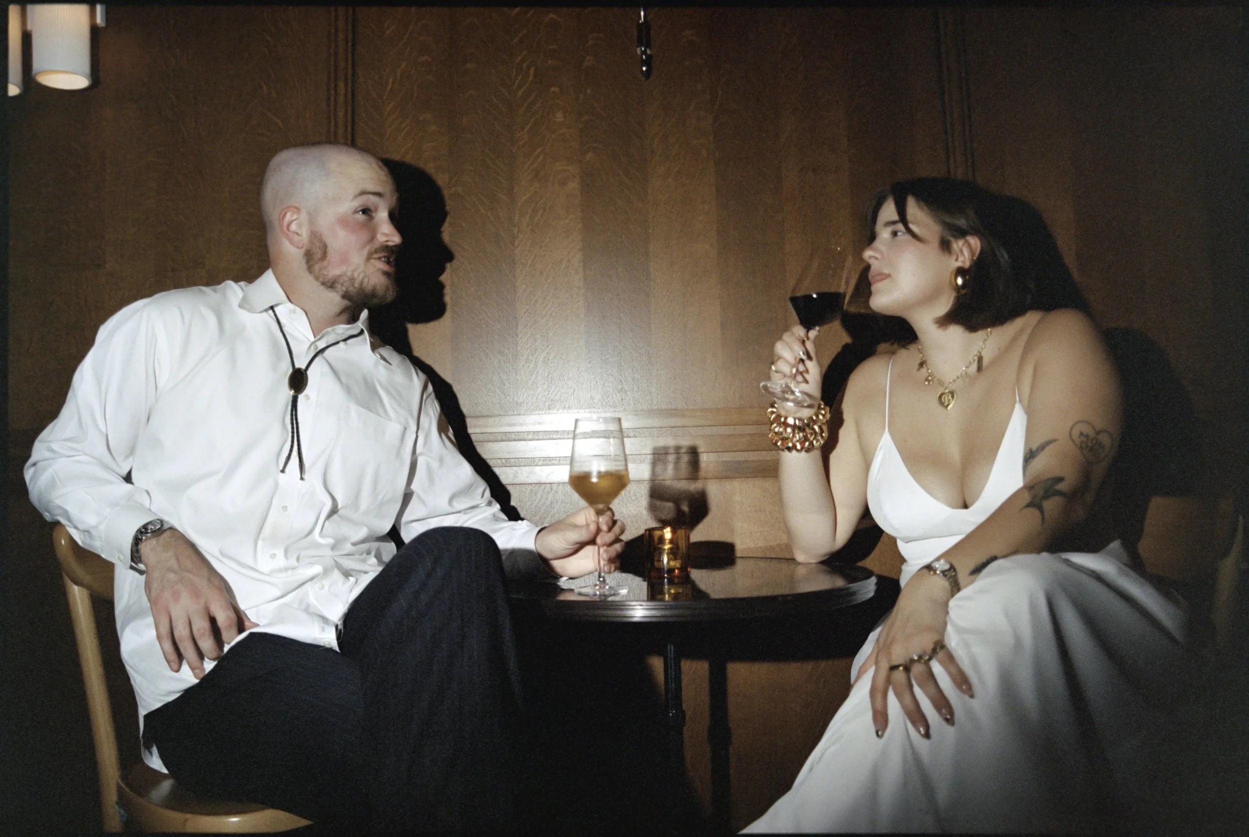 A man and woman sit at a table in a dimly lit restaurant, engaging in conversation. The woman is holding a glass of red wine and smiling, wearing a white dress and gold jewelry. The man is wearing a white shirt, listening attentively. Drinks are on the table, and the background has warm wooden paneling.