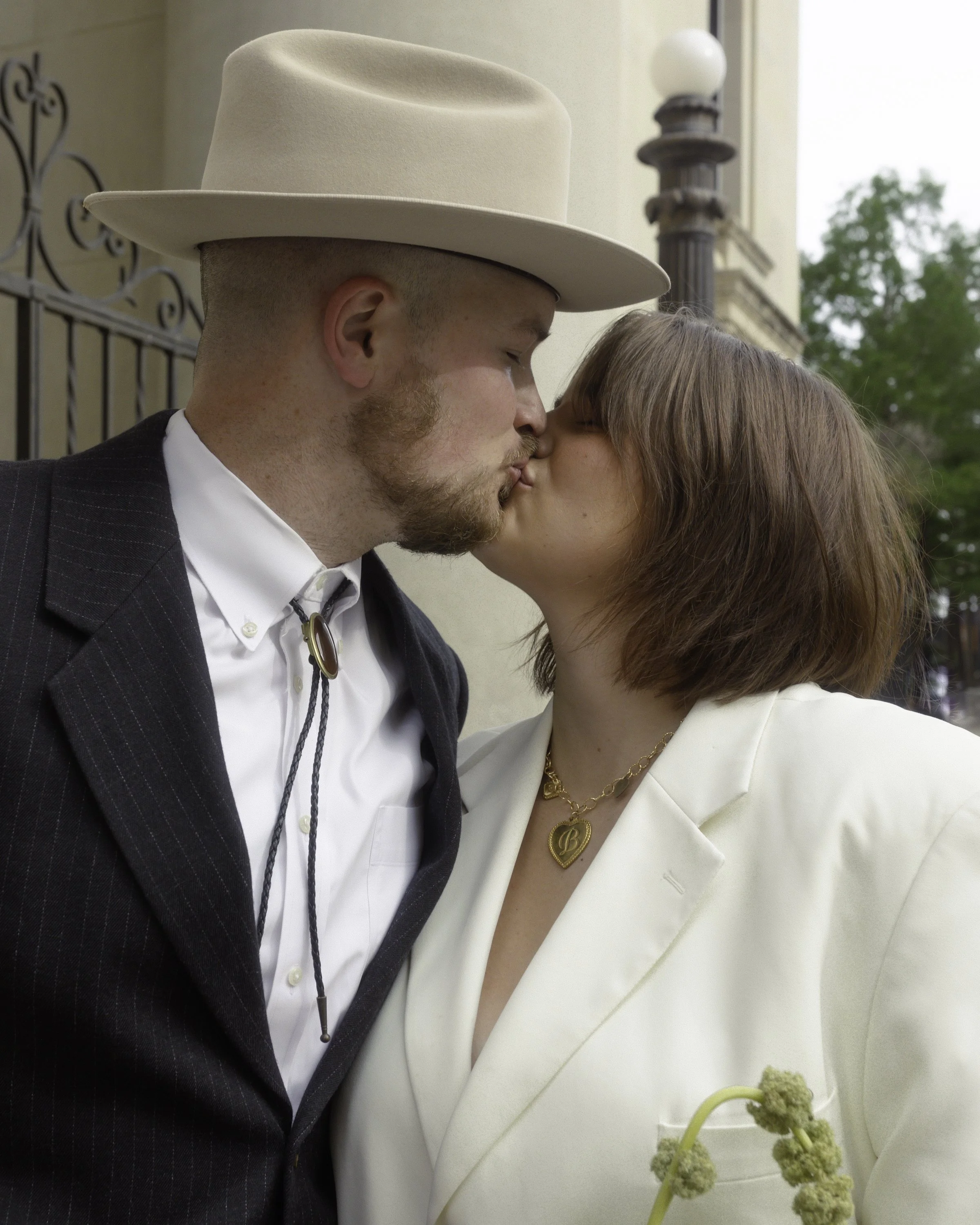 A man and woman kissing. The man is wearing a large cream hat and striped suit, and the woman is in a cream blazer with gold jewelry. Background includes a fence, lamp, and tree.