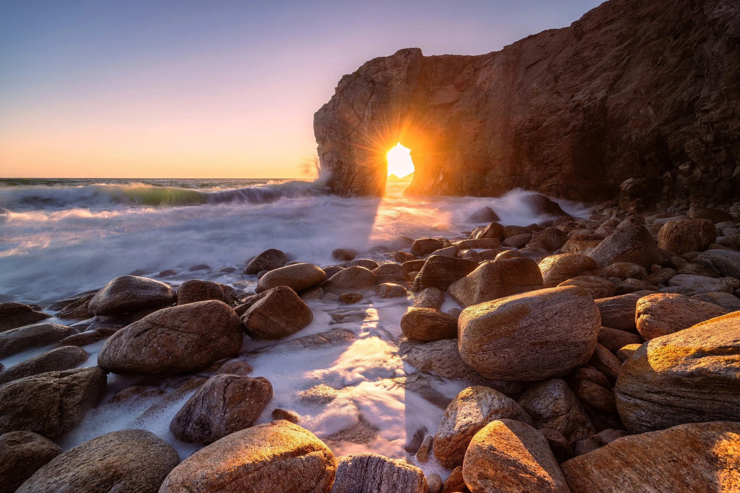 sun sets through natural hole in beach cliff waves rolling in on rocky shore