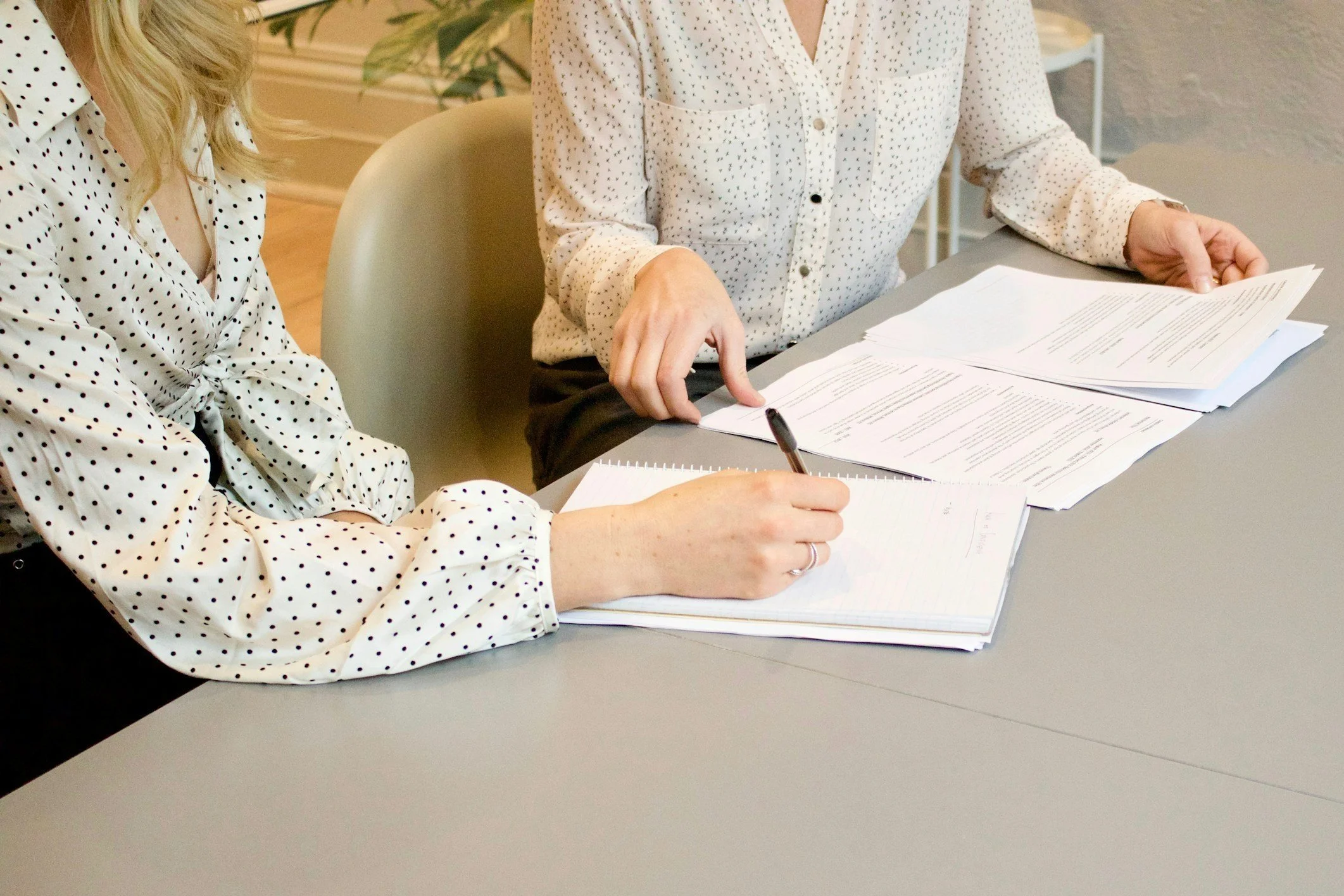Two women wearing white blouses with black polka dots are sitting at a gray table, reviewing and taking notes on documents and paper stacks.