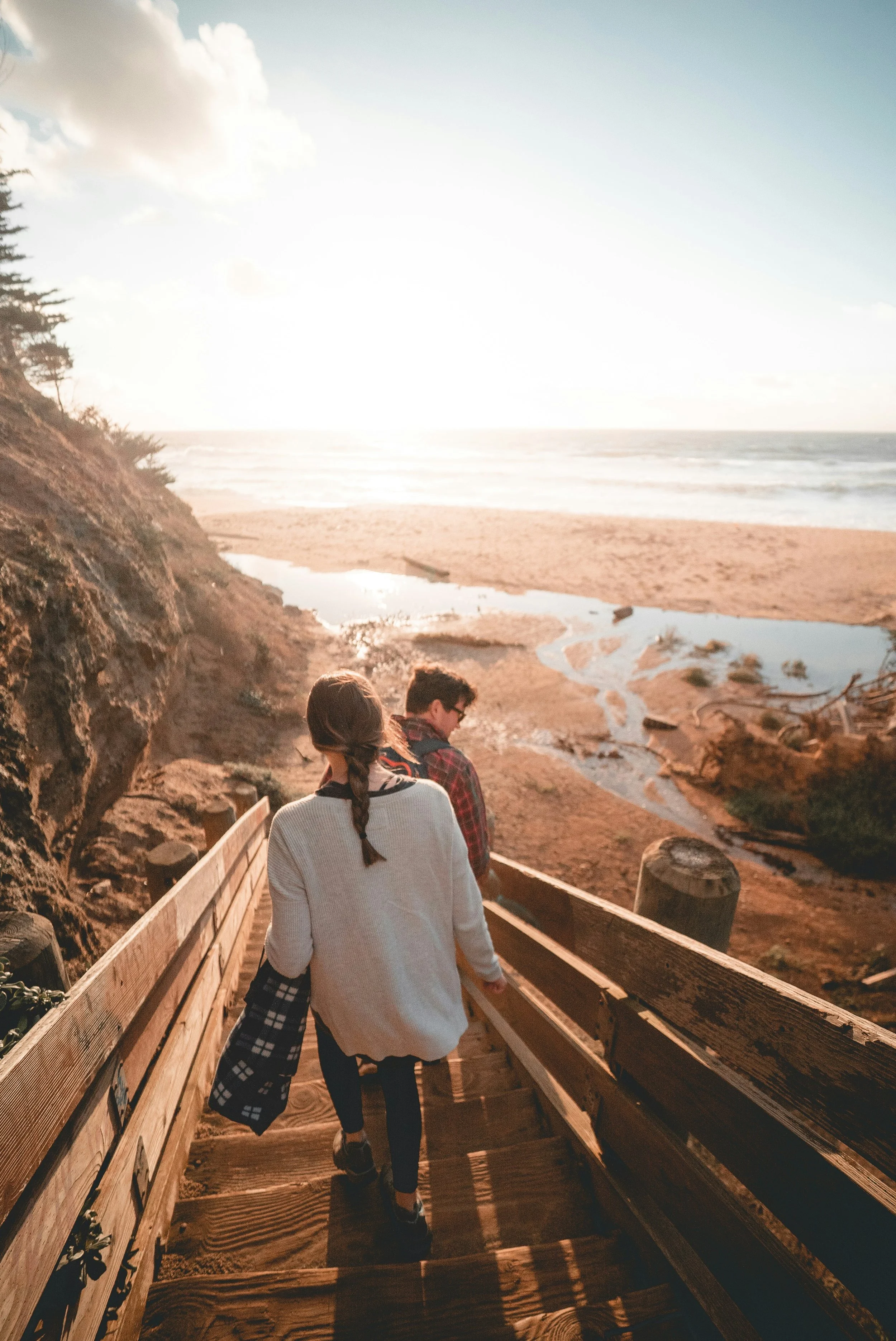 woman with braid and man with sunglasses walk down wooden stairs to sandy beach