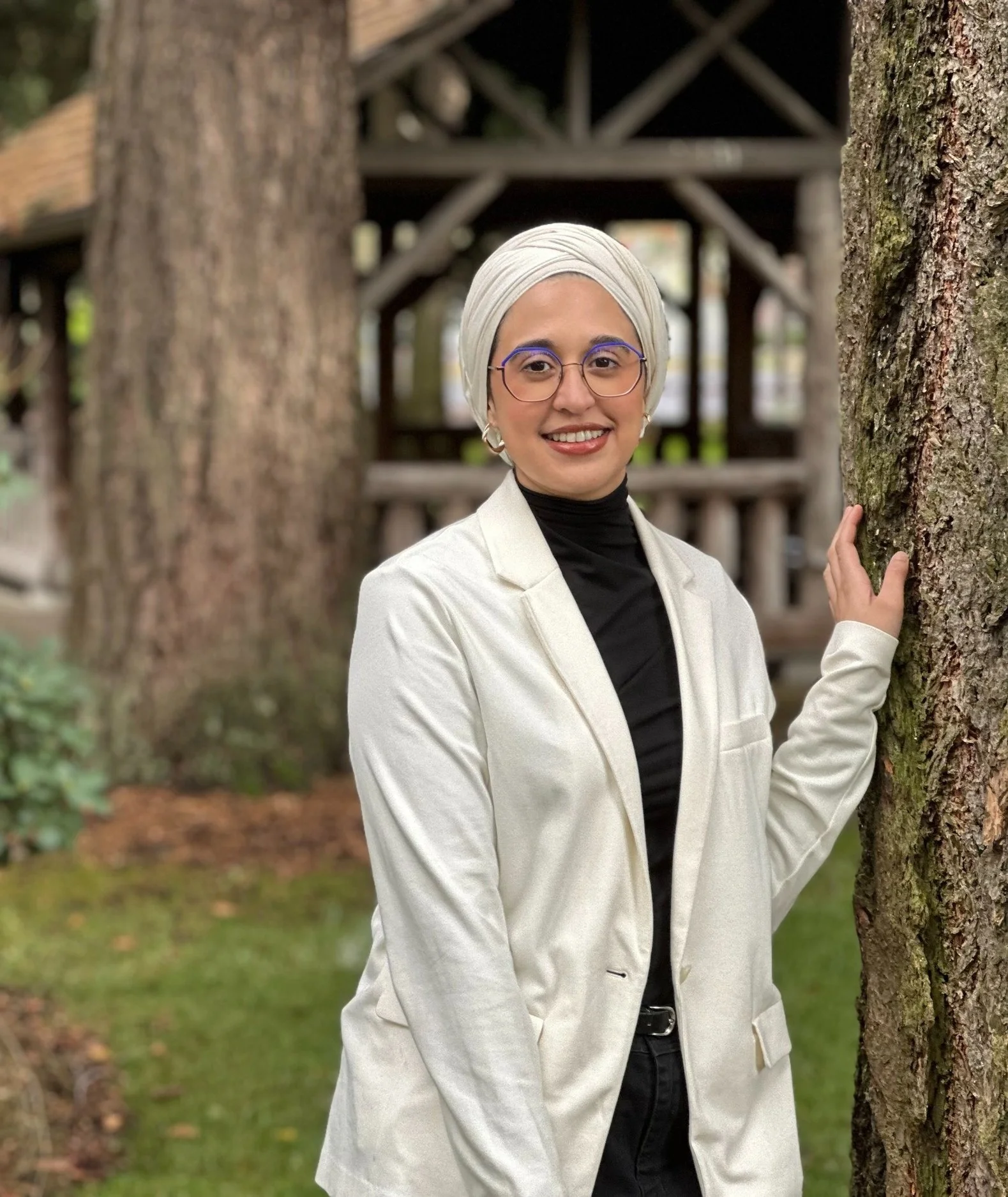 Image shows Dr. Faraha Hasan, clinical psychologist and post-doctoral fellow, standing with hand on tree, wearing white head covering and jacket