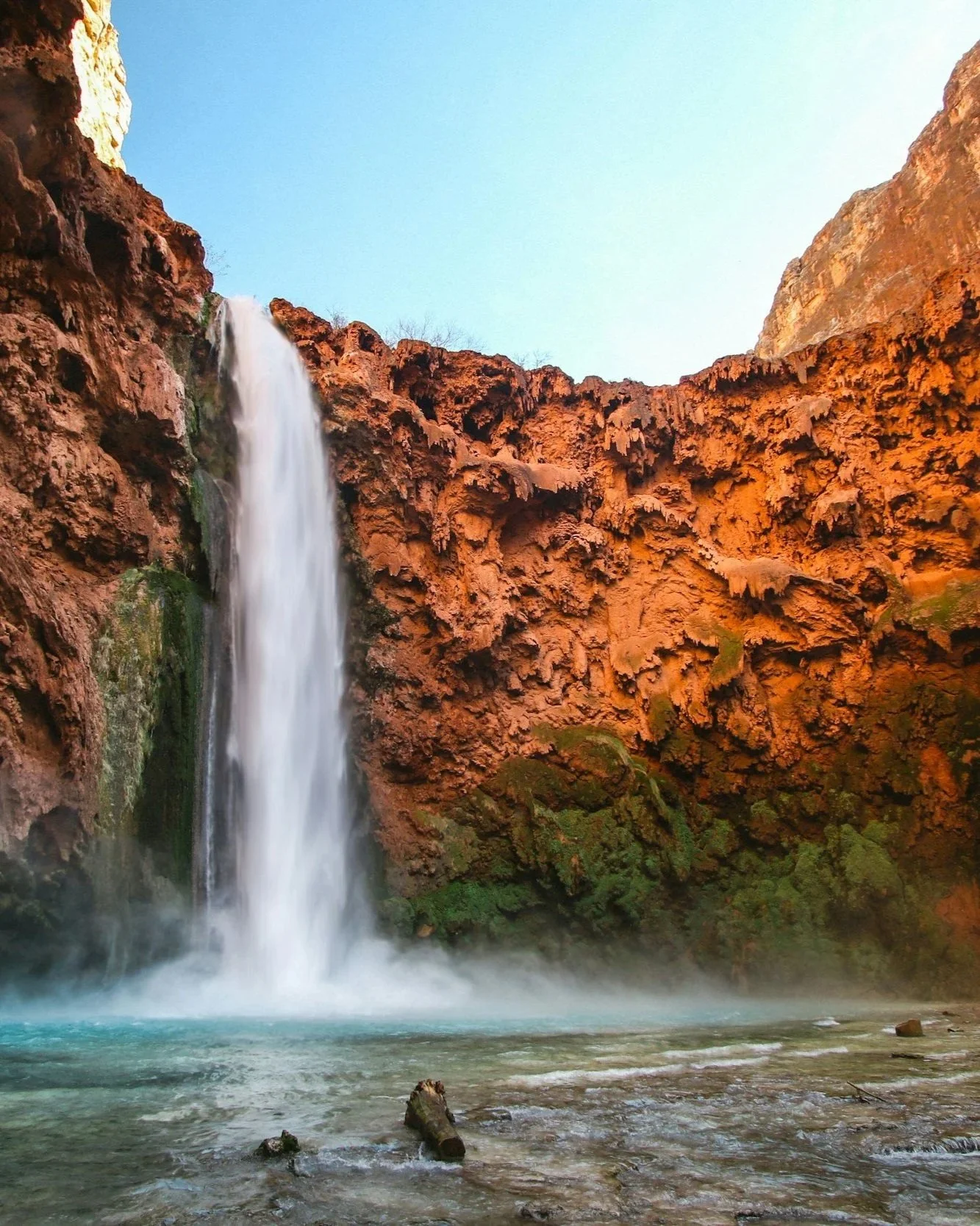 Photo of tall white waterfall cascading over red canyon cliff into cool blue and green pool below.