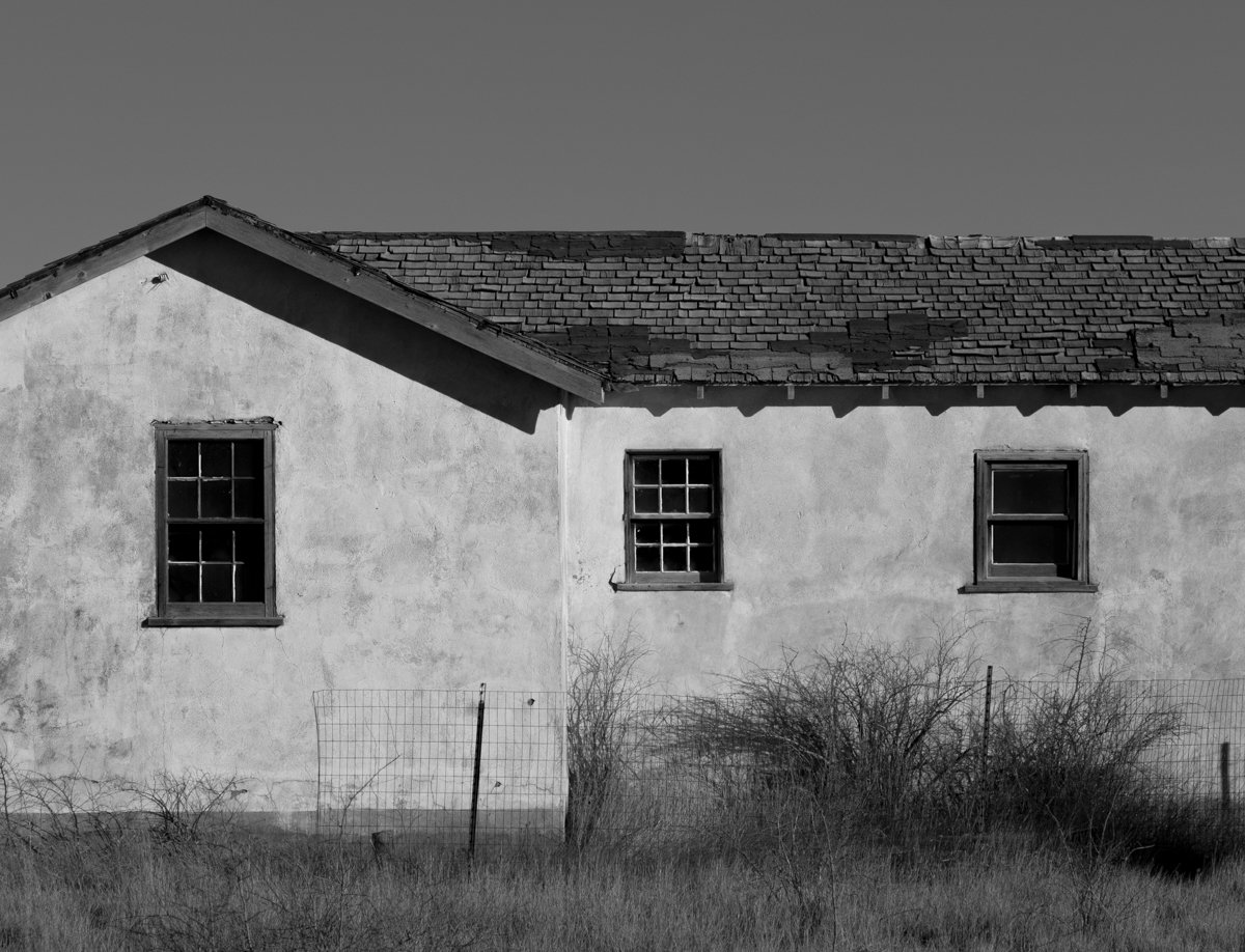 An old, abandoned house with a weathered exterior, three broken or missing window panes, and a roof with damaged shingles. Overgrown weeds and a wire fence are in the foreground.