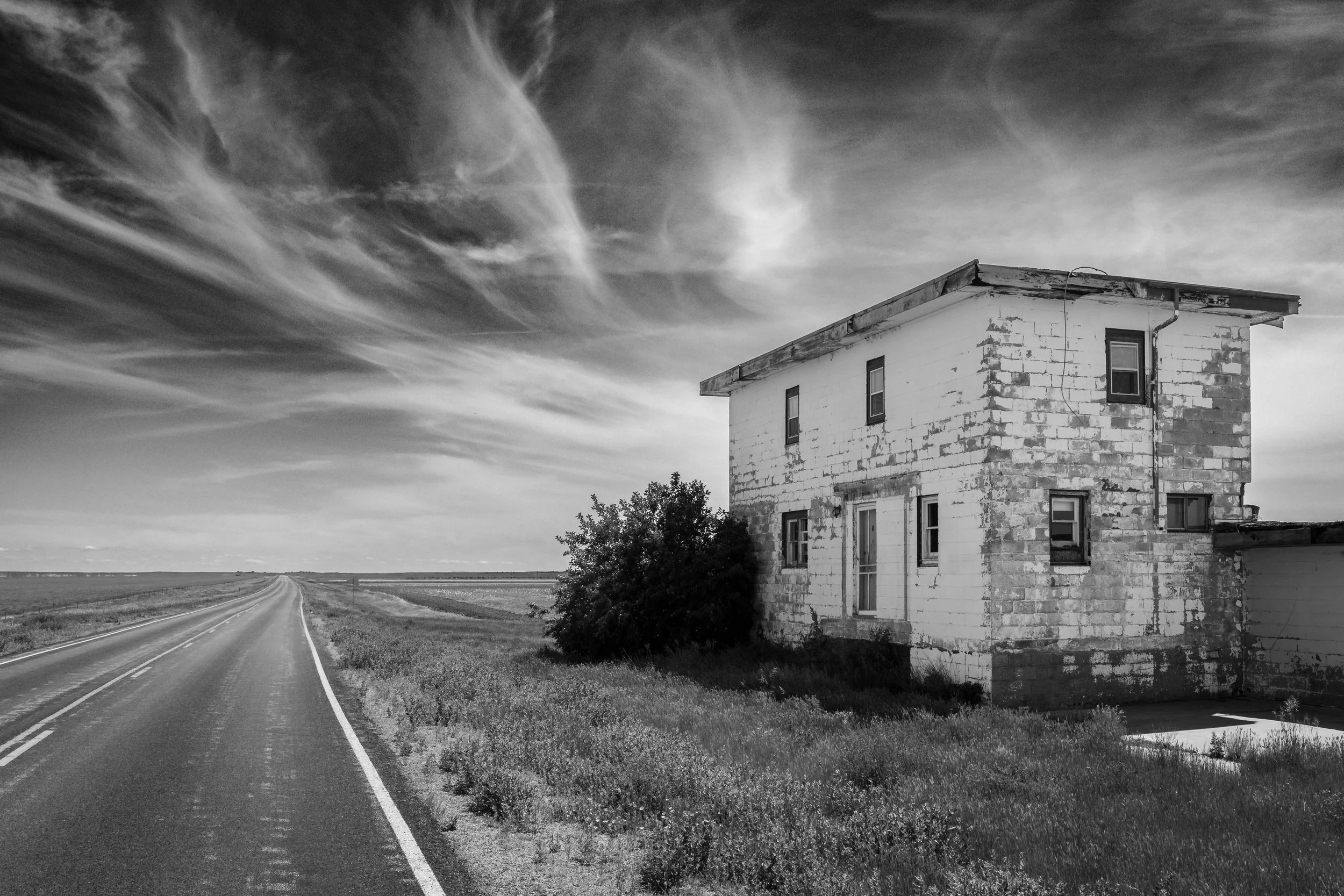 Black and white photo of a two-story house made of concrete blocks, with a slanted roof, on the right side. A narrow, straight road stretches into the distance on the left, surrounded by grassy fields and a few trees. The sky is filled with wispy clouds.