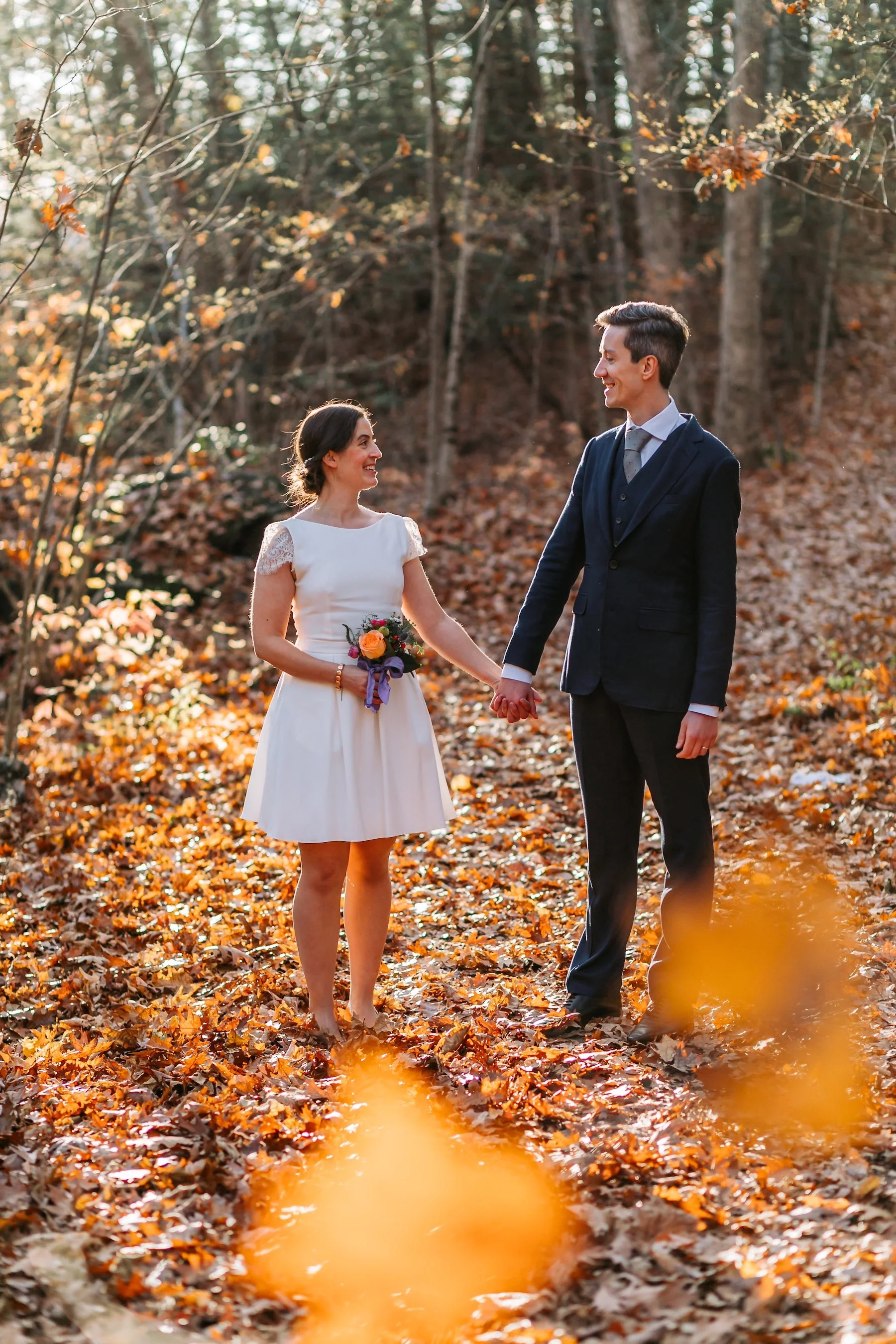Bride and groom walking hand in hand along a woodland path covered in autumn leaves.