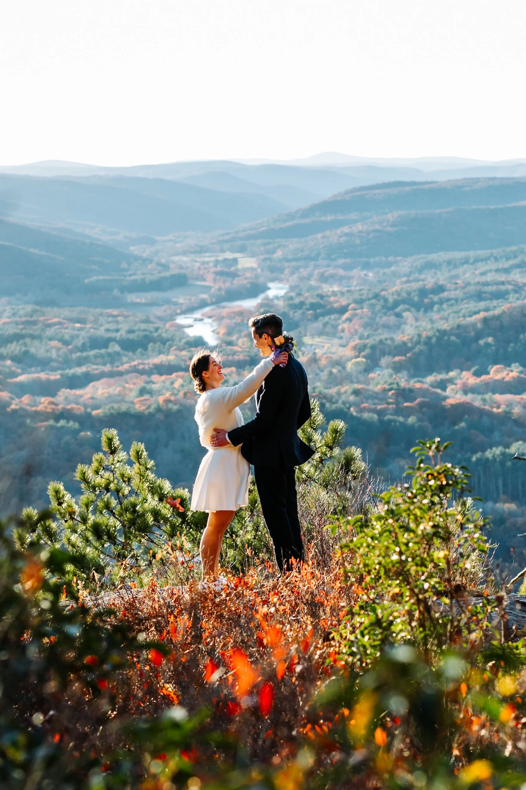 Couple in a white wedding dress and navy suit sharing a smile at a mountain overlook surrounded by New England fall foliage.