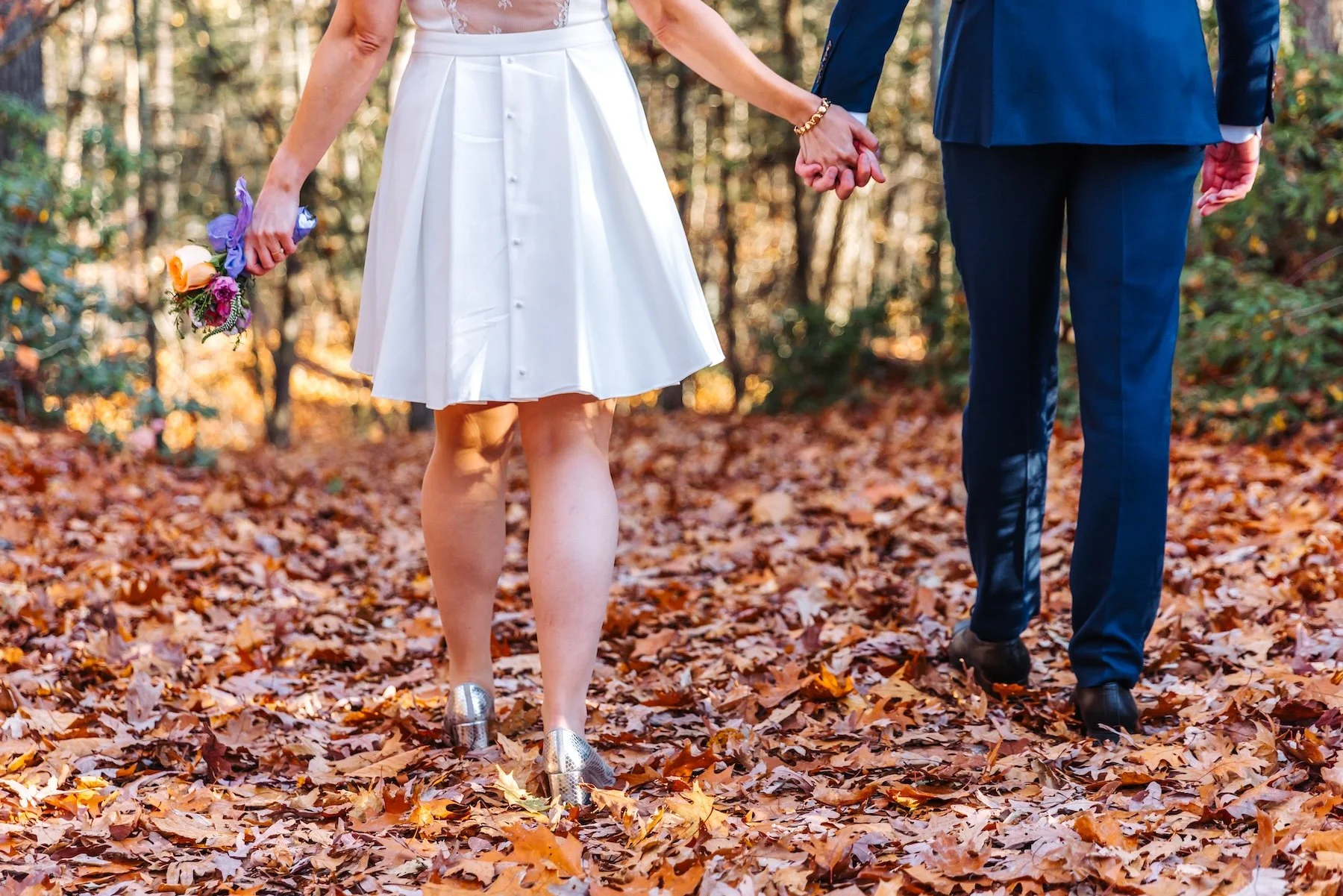 Newlyweds holding hands on a woodland trail covered in fallen autumn leaves during an intimate Massachusetts elopement.