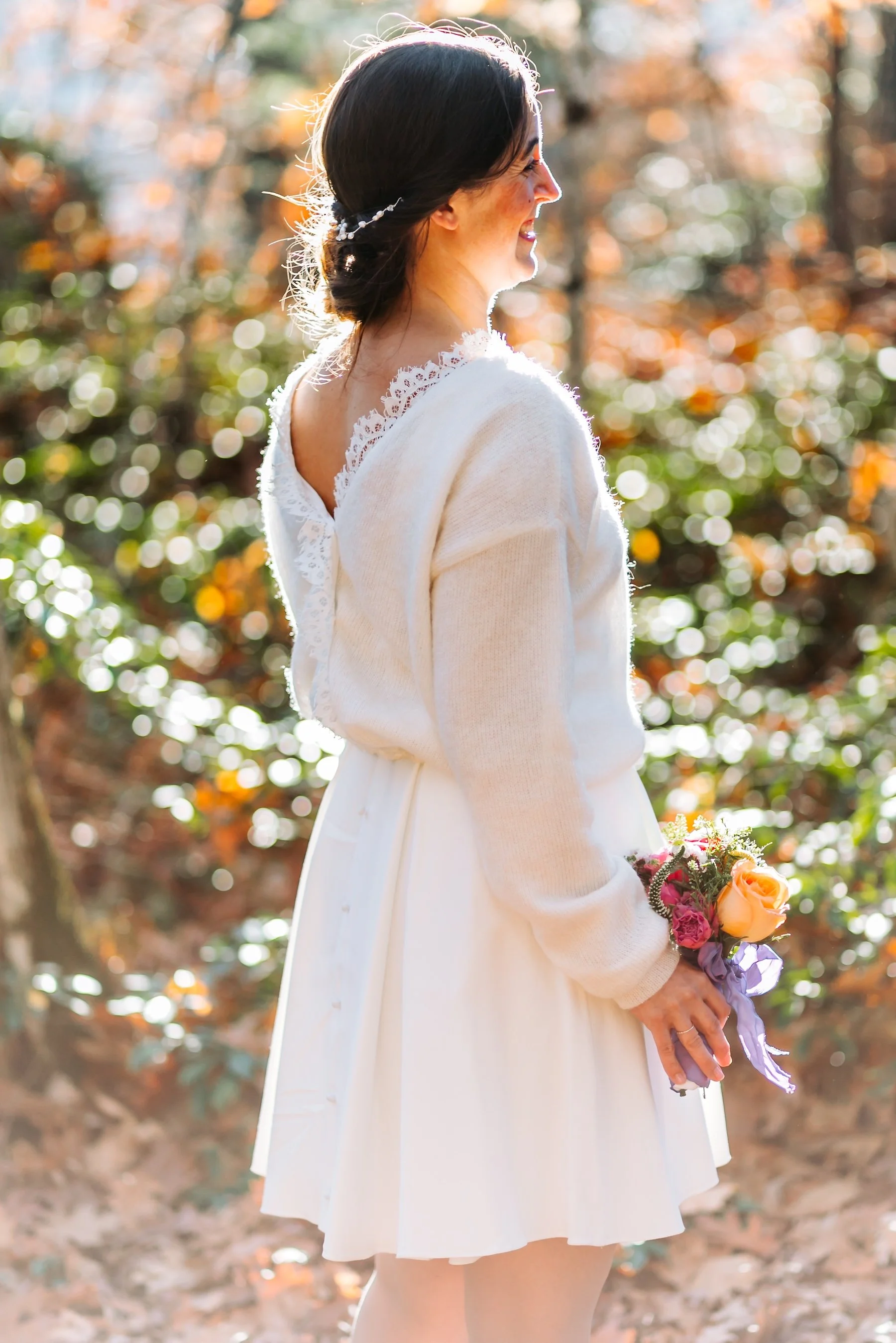 Bride in a white dress and cream sweater holding a colorful bouquet in the warm afternoon light of an autumn forest.