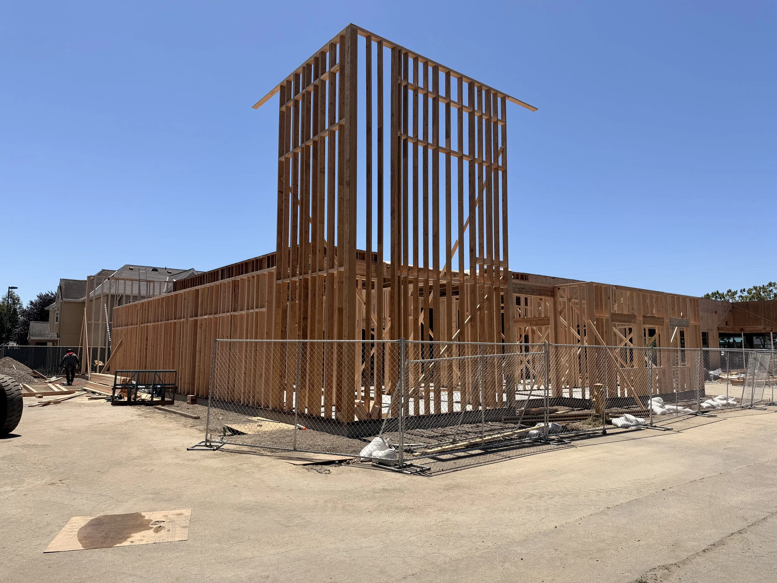 Construction site with a wooden framework of a building under development, surrounded by temporary fencing, with nearby houses and a clear blue sky.