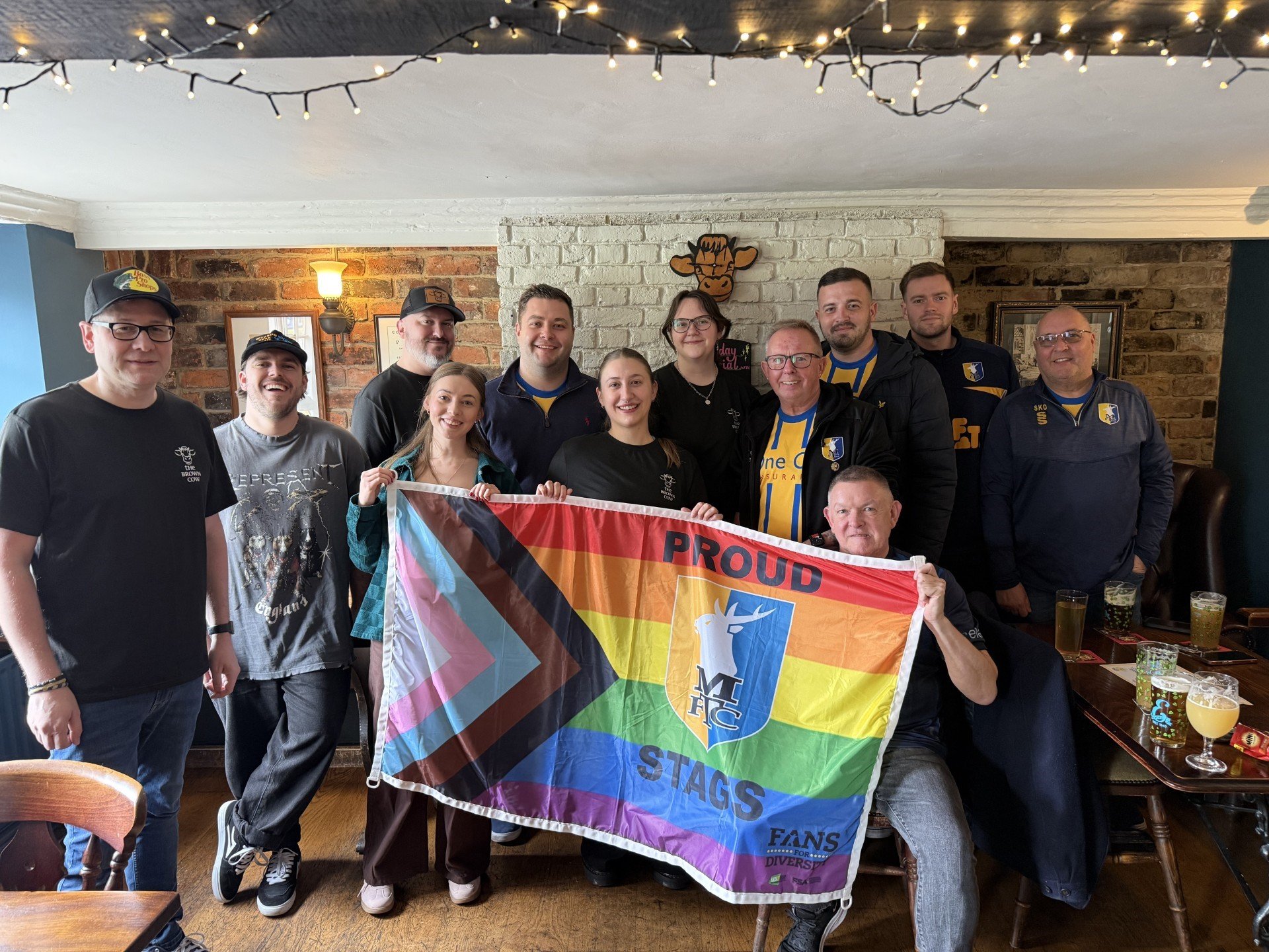 Members from Proud Stags, and staff from the Brown Cow, pose for a group picture, with the Proud Stags flag being held up in front of everyone.