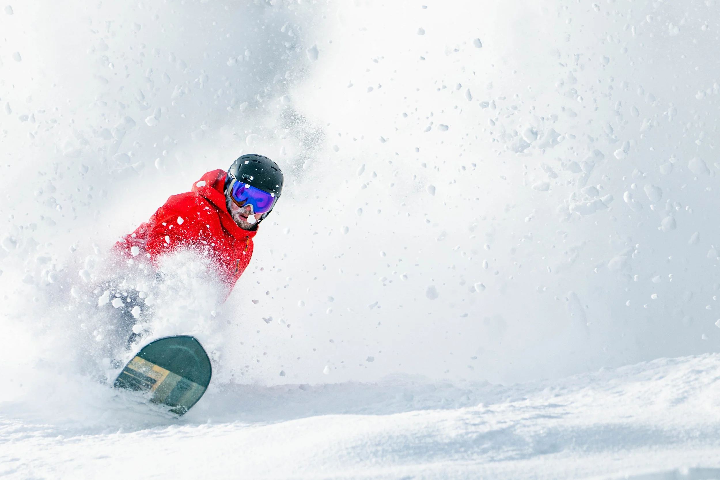 A snowboarder in a red jacket and black helmet carving through fresh snow, creating a spray of snowflakes in the air.