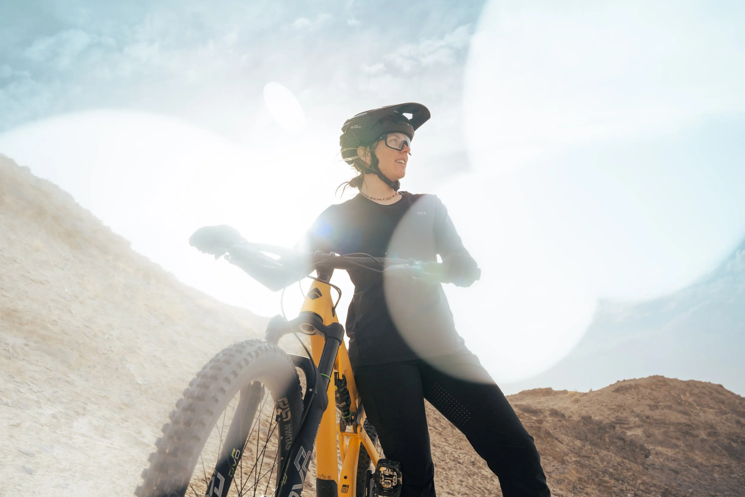 A woman with a bicycle, wearing a helmet and sunglasses, standing on a rugged terrain with mountains in the background.
