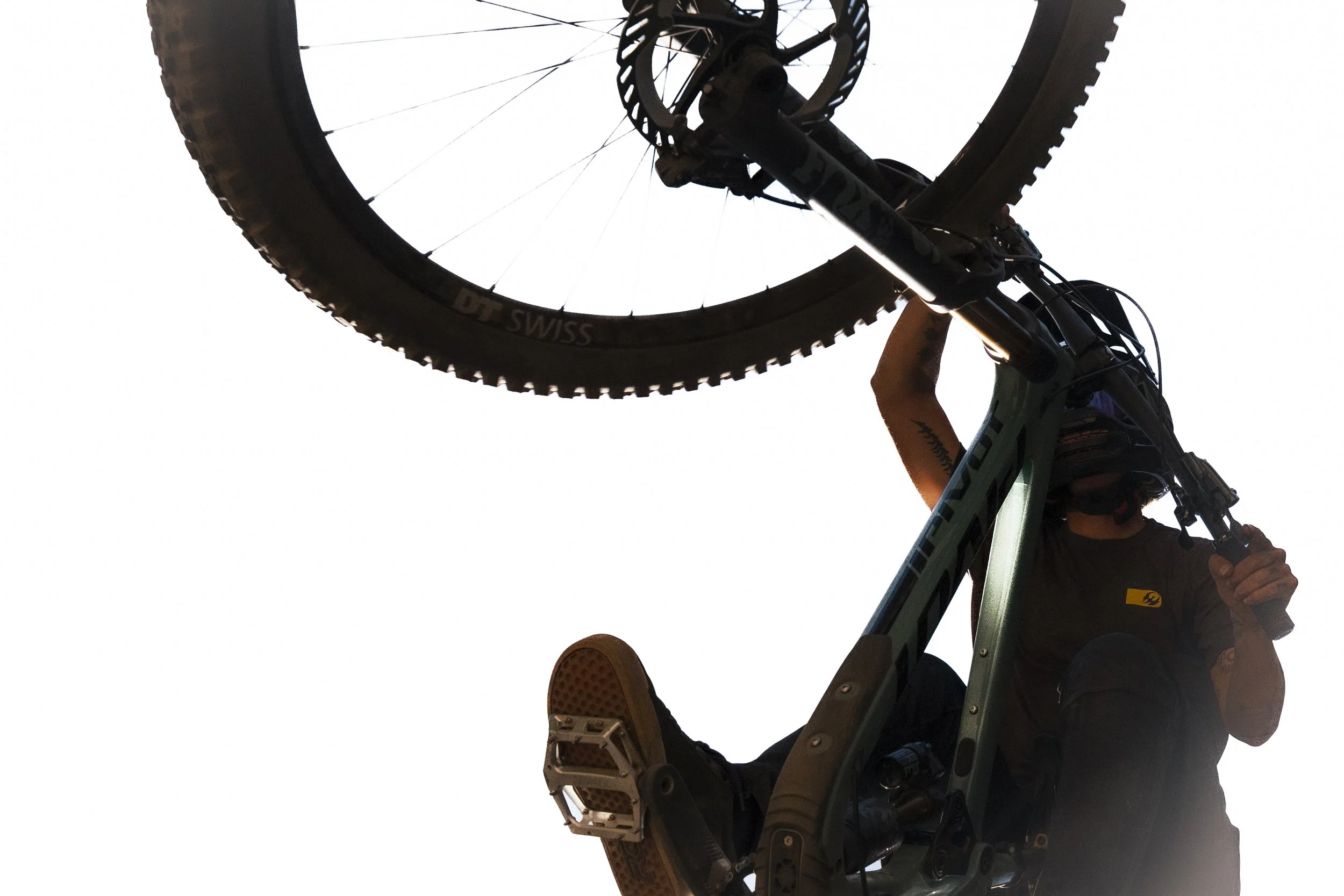 A person wearing a helmet and black clothing is holding a mountain bike above their head, with the bike's front wheel and parts of the frame visible against a bright sky.