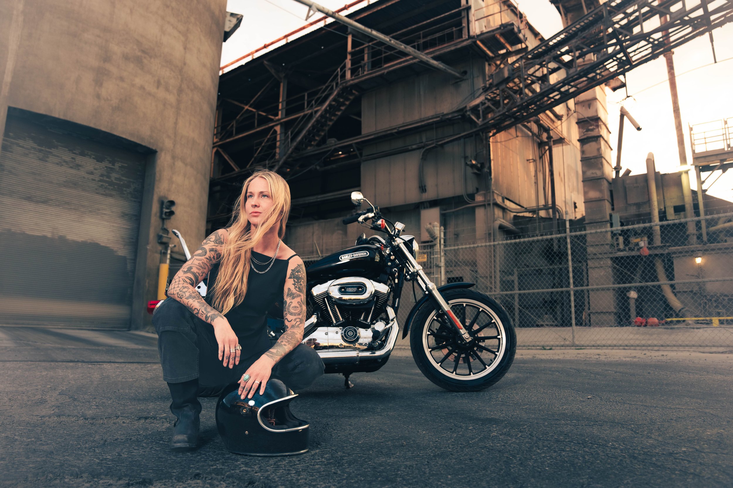 A woman with long blonde hair, tattoos on her arms, and wearing a black shirt, black pants, and black boots, crouching next to a black Harley-Davidson motorcycle on an urban street. She has a black motorcycle helmet on the ground and is holding a cha