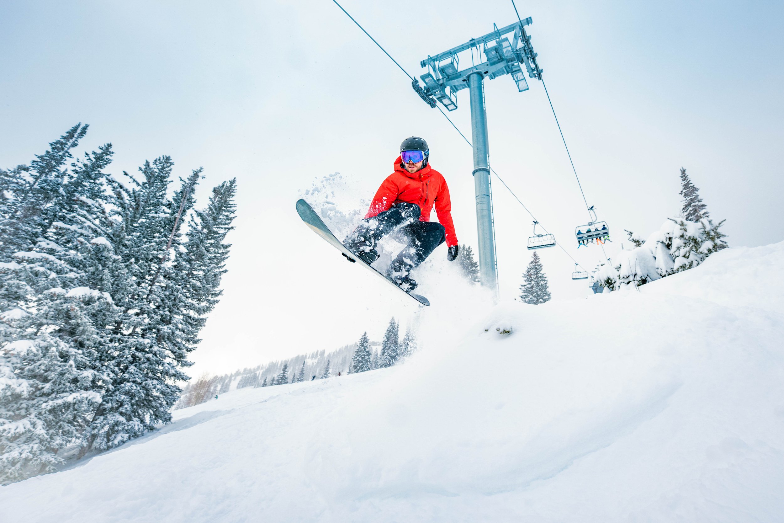 A person wearing a red jacket, black pants, and a black helmet snowboarding down a snowy slope near a ski lift, with snow-covered trees in the background.