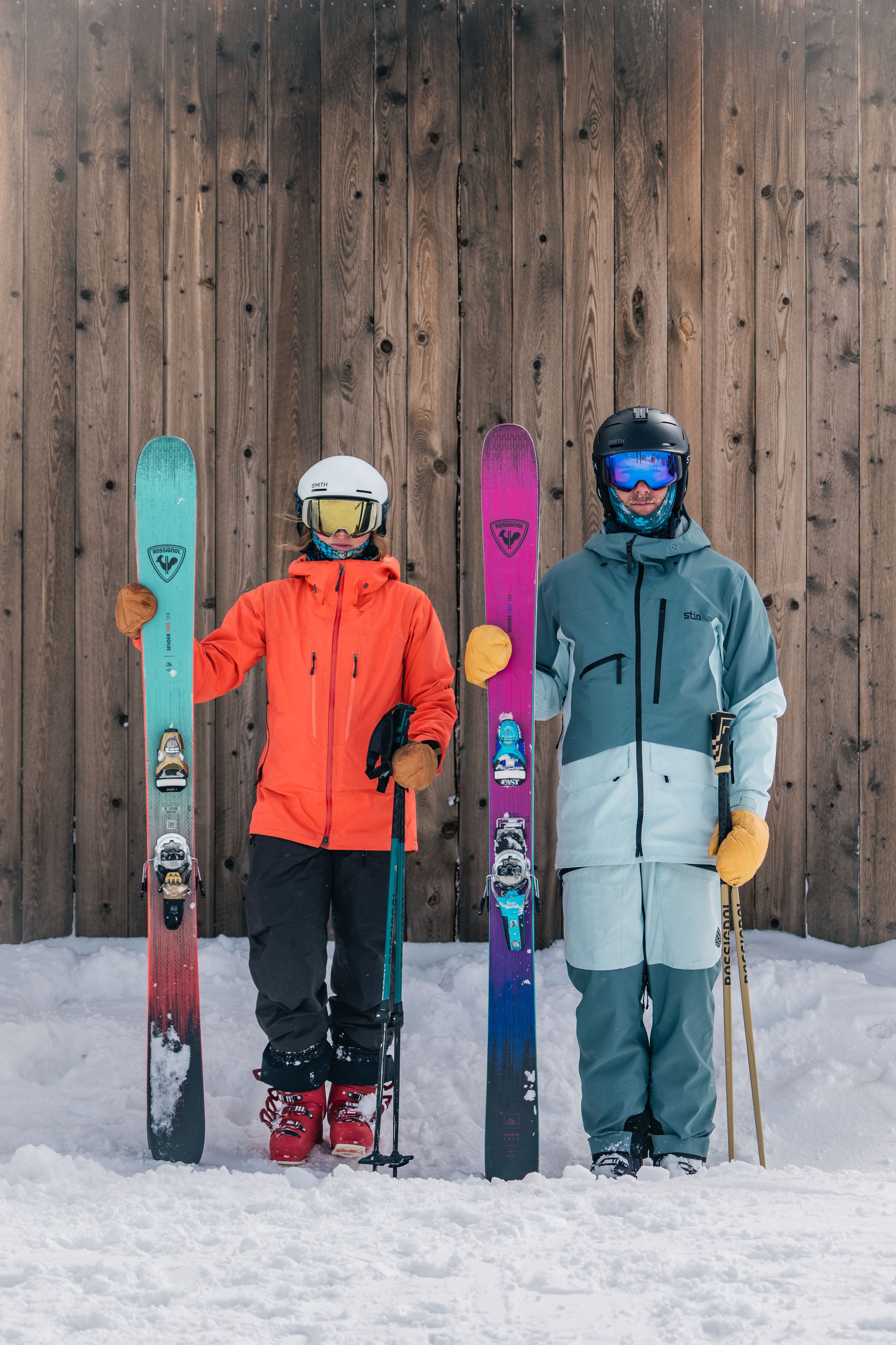 Two skiers standing on snow in front of a wooden wall, wearing helmets, goggles, winter jackets, pants, boots, and holding skis and poles.