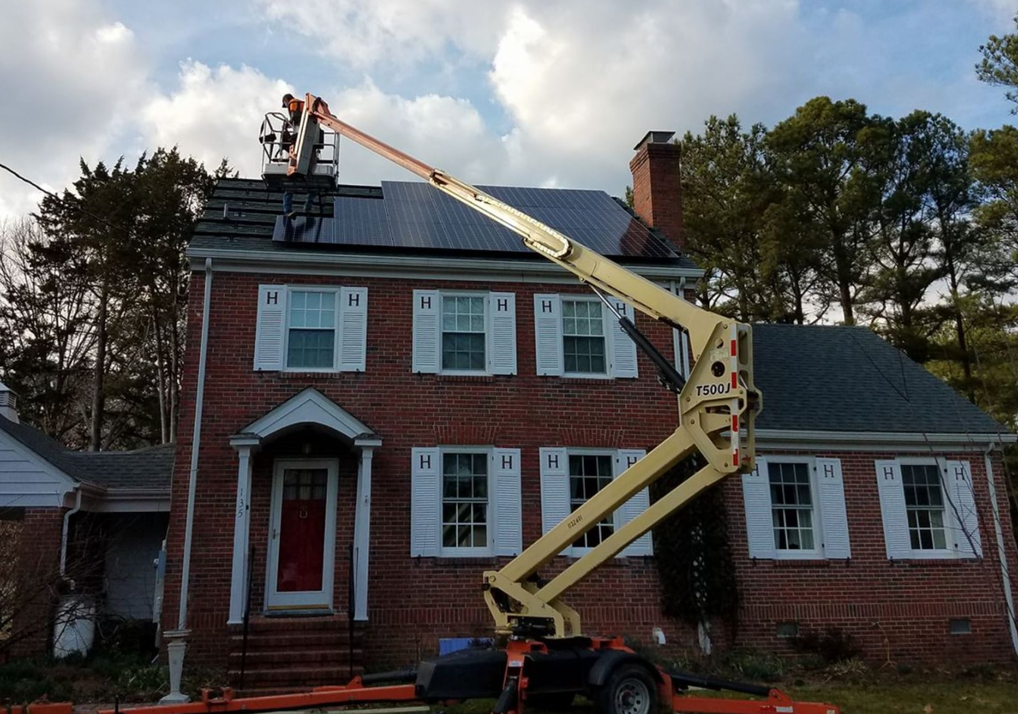 A person installing solar panels on the roof of a two-story brick house using a lift.