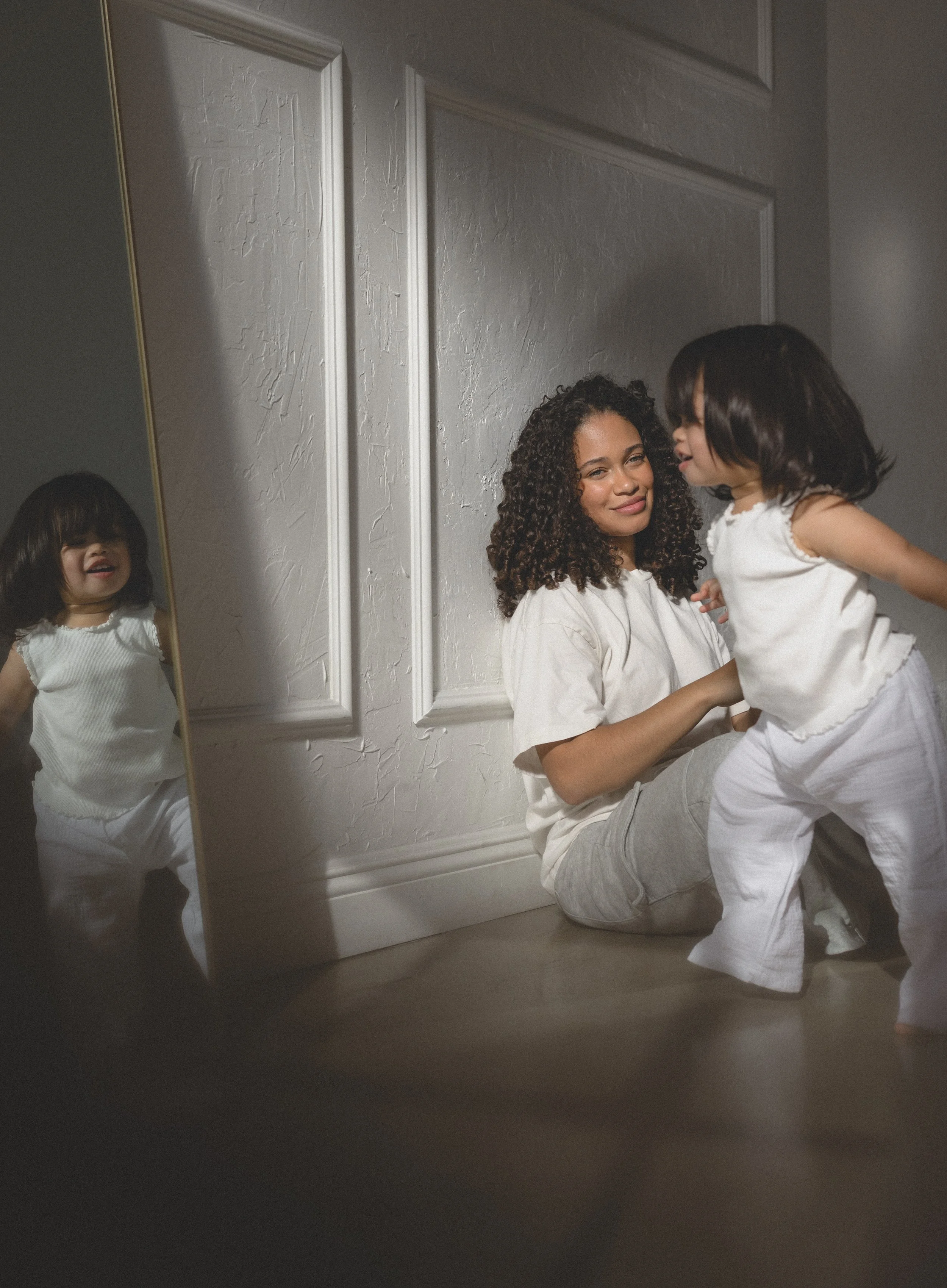A woman with curly hair is sitting on the floor, smiling, while a girl with straight hair is standing in front of her. Their reflections are seen in a mirror on the left side, and the girl is slightly blurred.