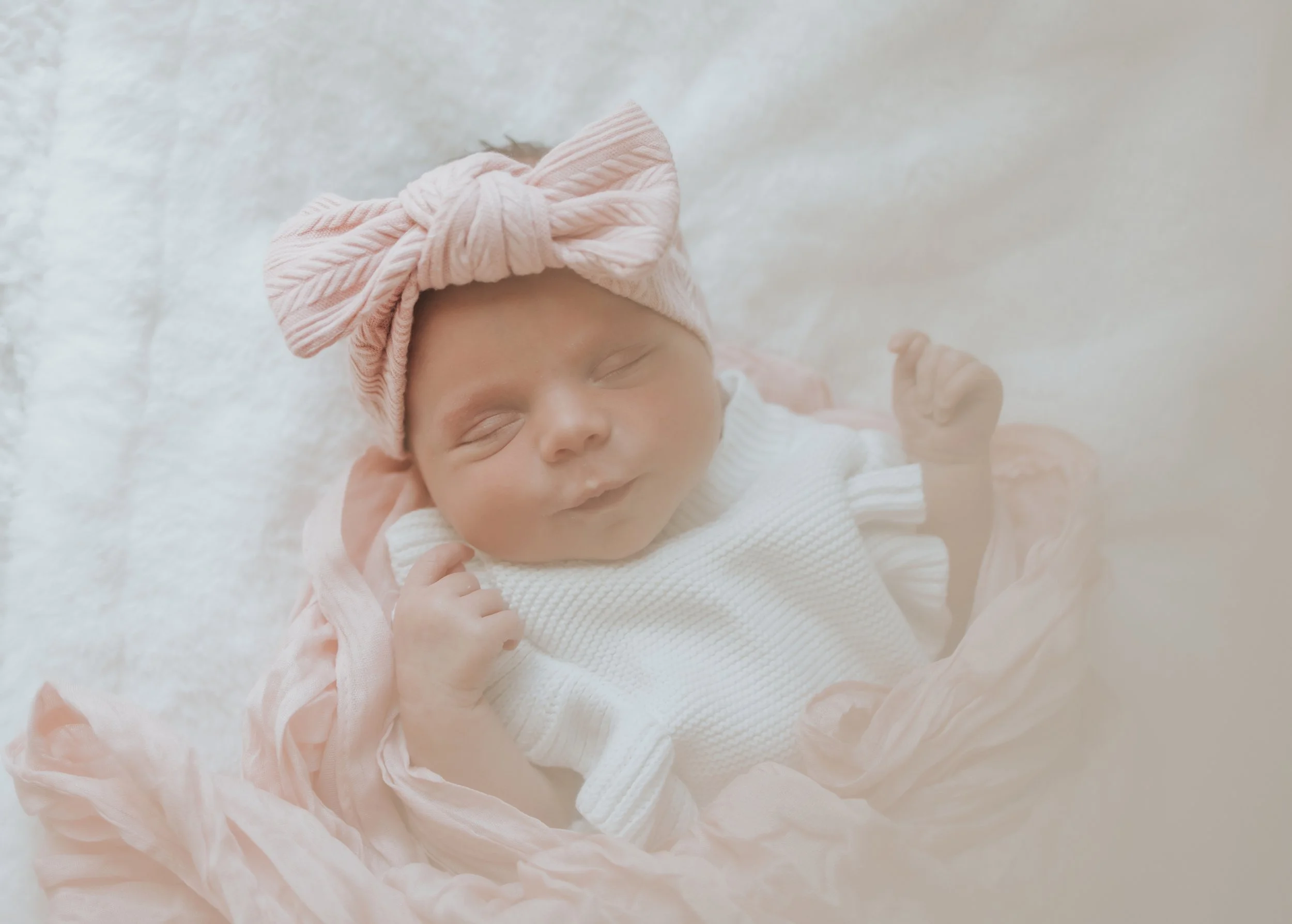 A sleeping baby girl wearing a pink headband with a bow and a white sweater, lying on a soft, fluffy white blanket.