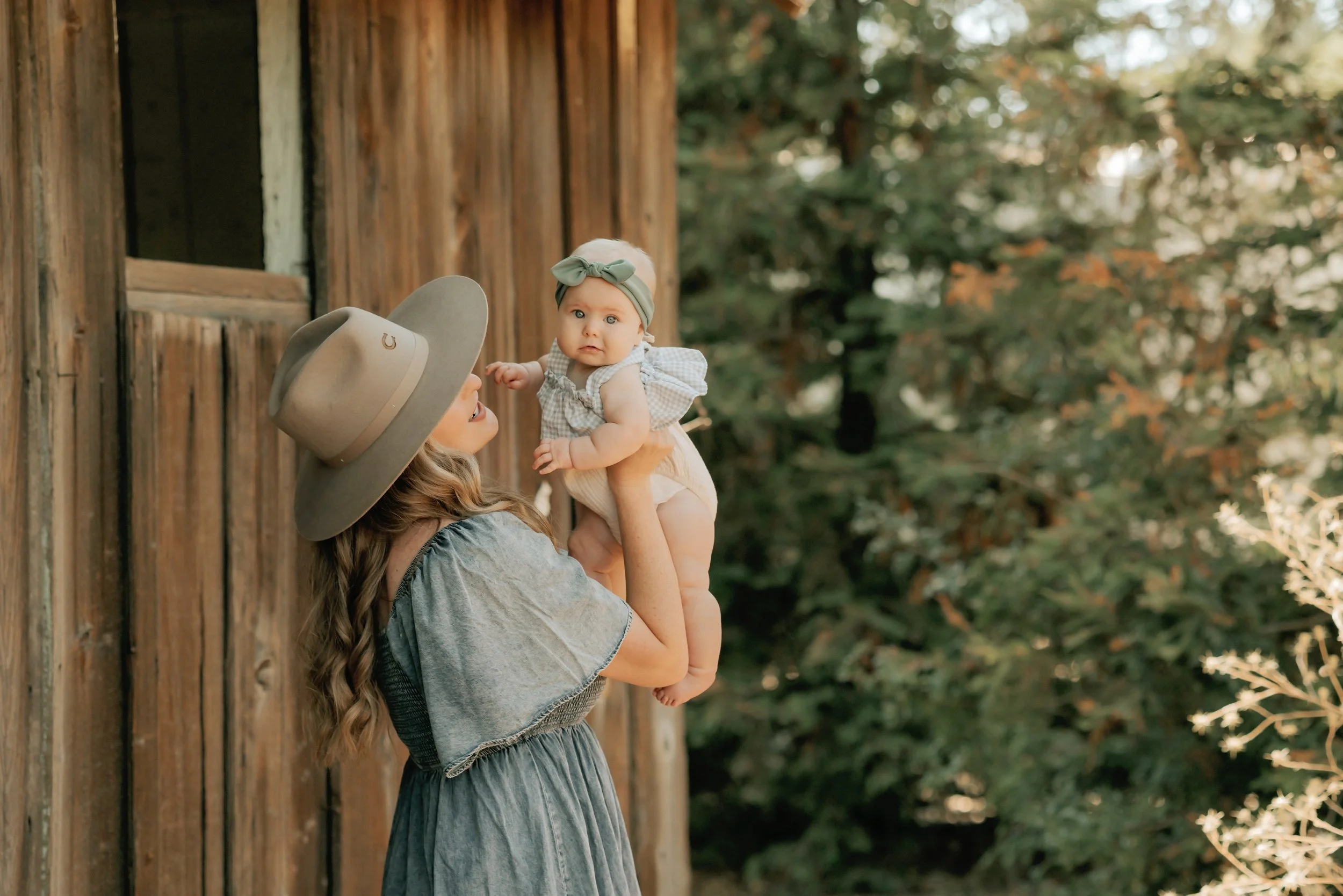 Woman holding baby near rustic wooden building and trees.