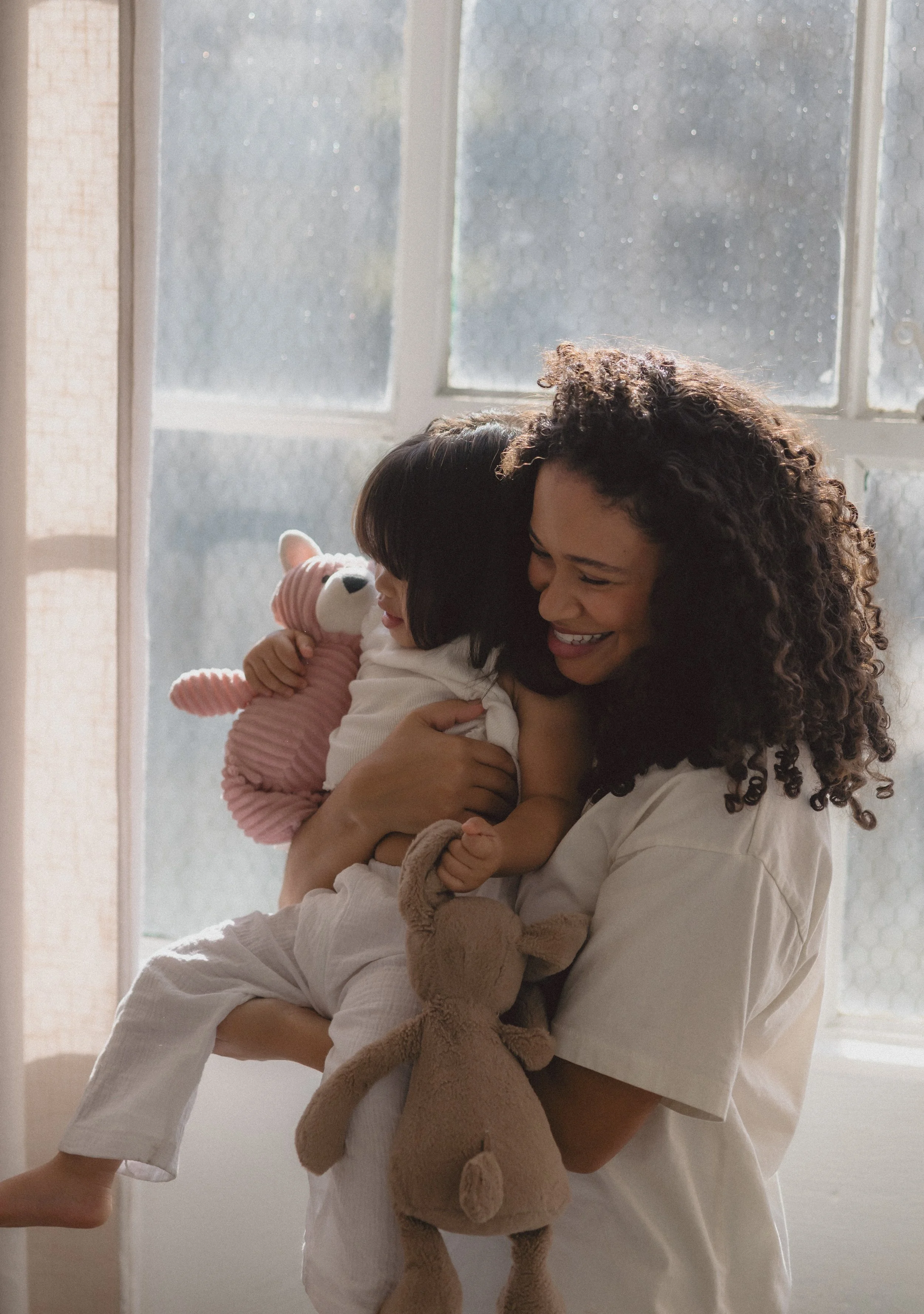 A woman holding a young girl with dark hair and a white shirt, both smiling. The girl is holding a pink stuffed animal, and the woman has a beige stuffed animal hanging from her arm. They are near a window with frosted glass, letting in soft natural light.