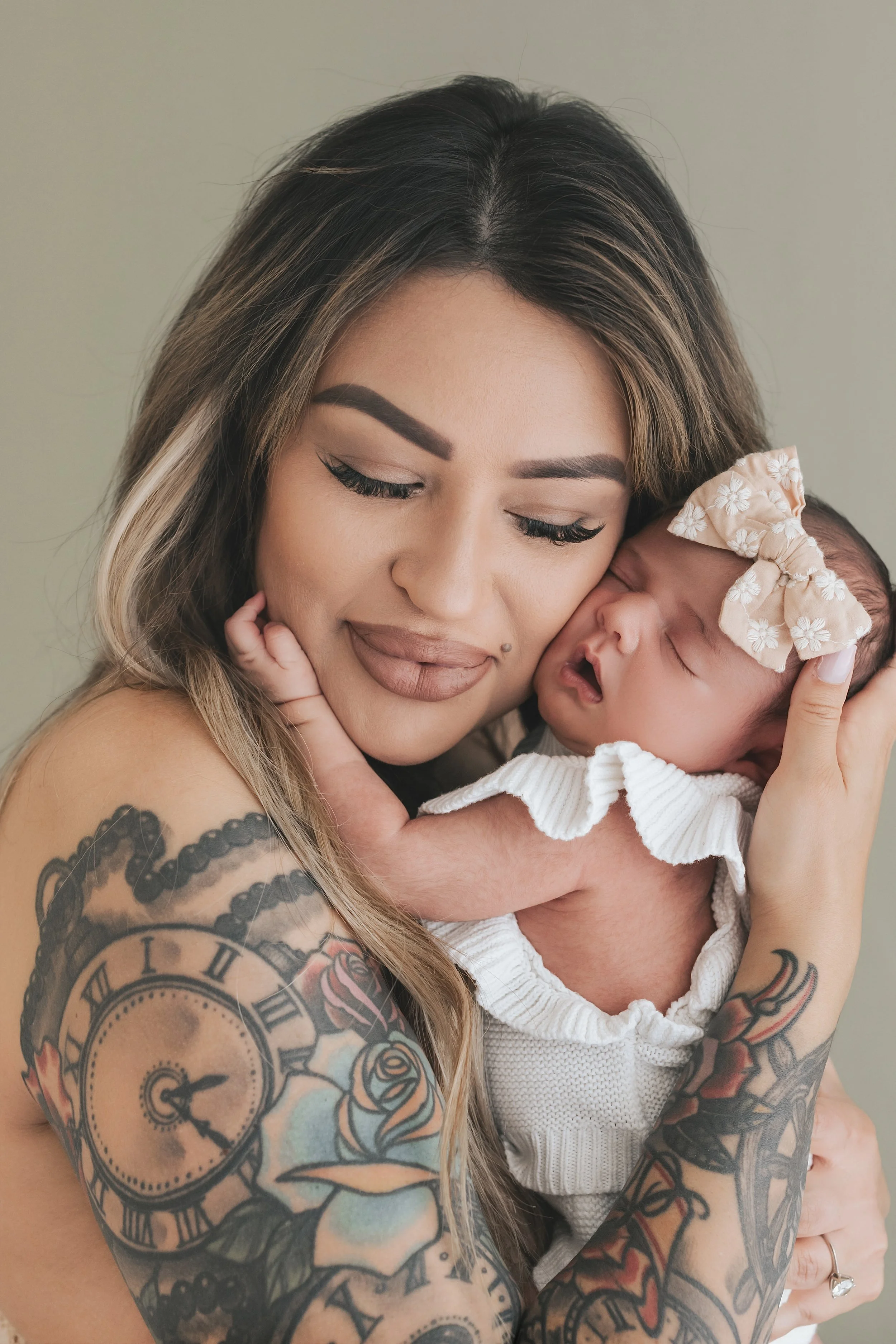 A woman with tattoos holding a newborn baby close to her face. The woman has long hair, well-defined eyebrows, and makeup. The baby is wearing a white outfit and a beige headband with a bow.