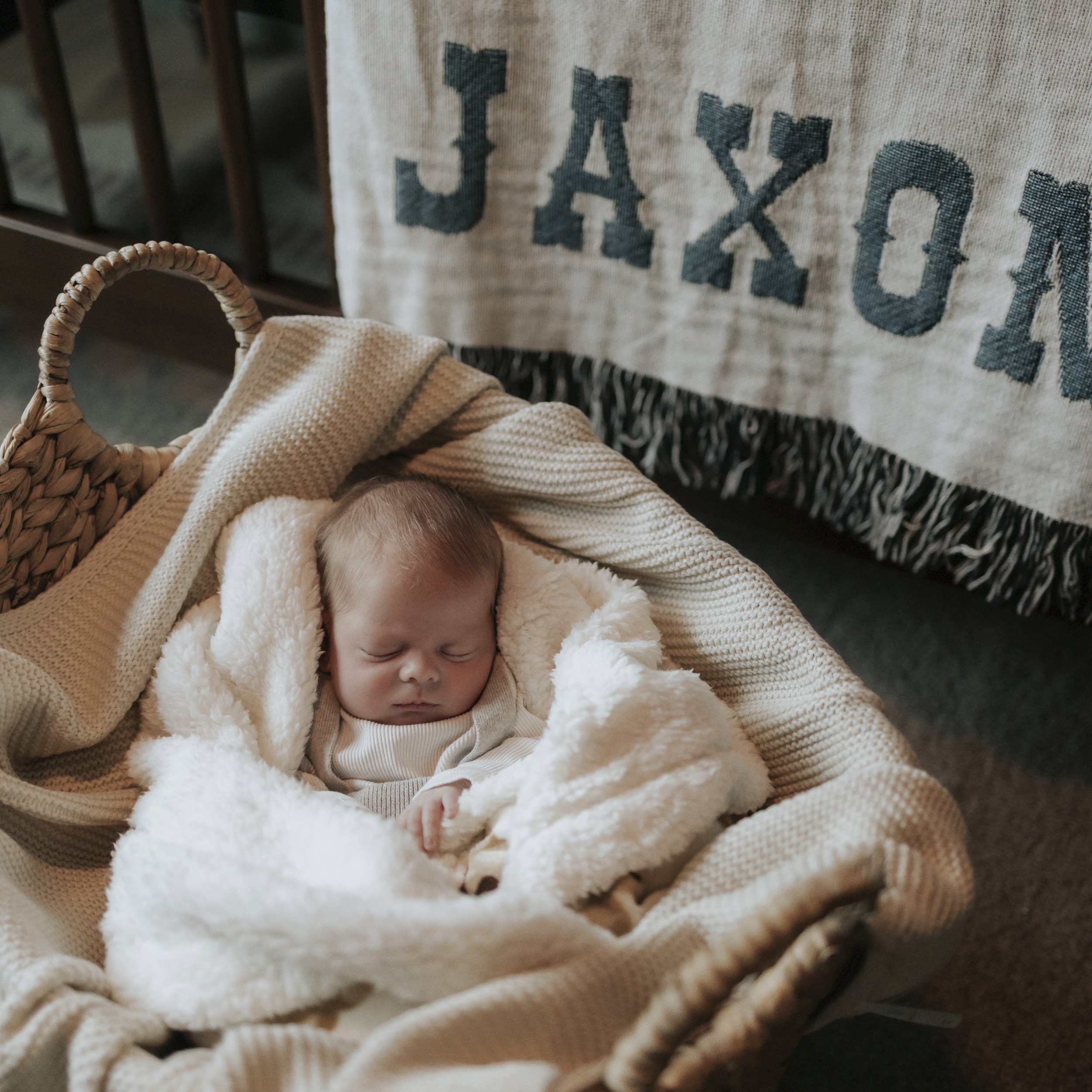 A sleeping baby in a woven bassinet, wrapped in a soft cream blanket, with a towel that has the word 'JAXON' hanging in the background.