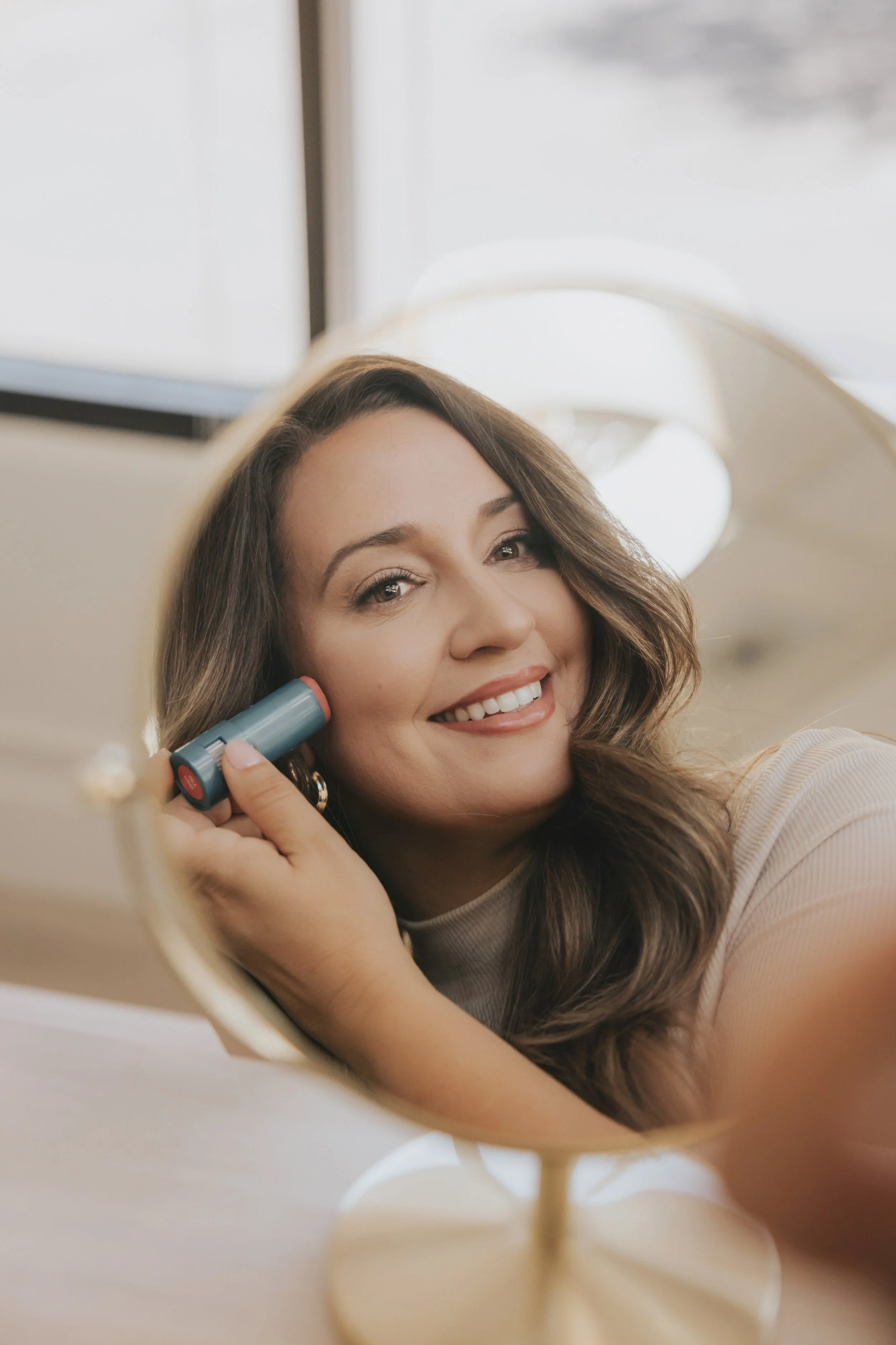 Woman smiling at her reflection in a mirror, applying lipstick with a lipstick tube.