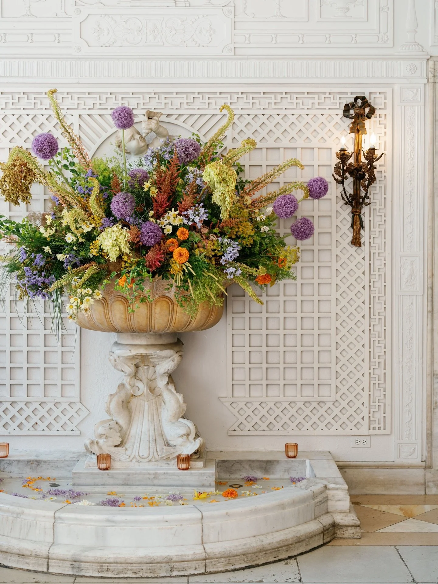 Sometimes, all you need is storied architecture and an unapologetically abundant composition.

Set against carved stone and timeworn detail, this arrangement leans into movement, texture, and a painterly palette, where sculptural alliums, trailing gr