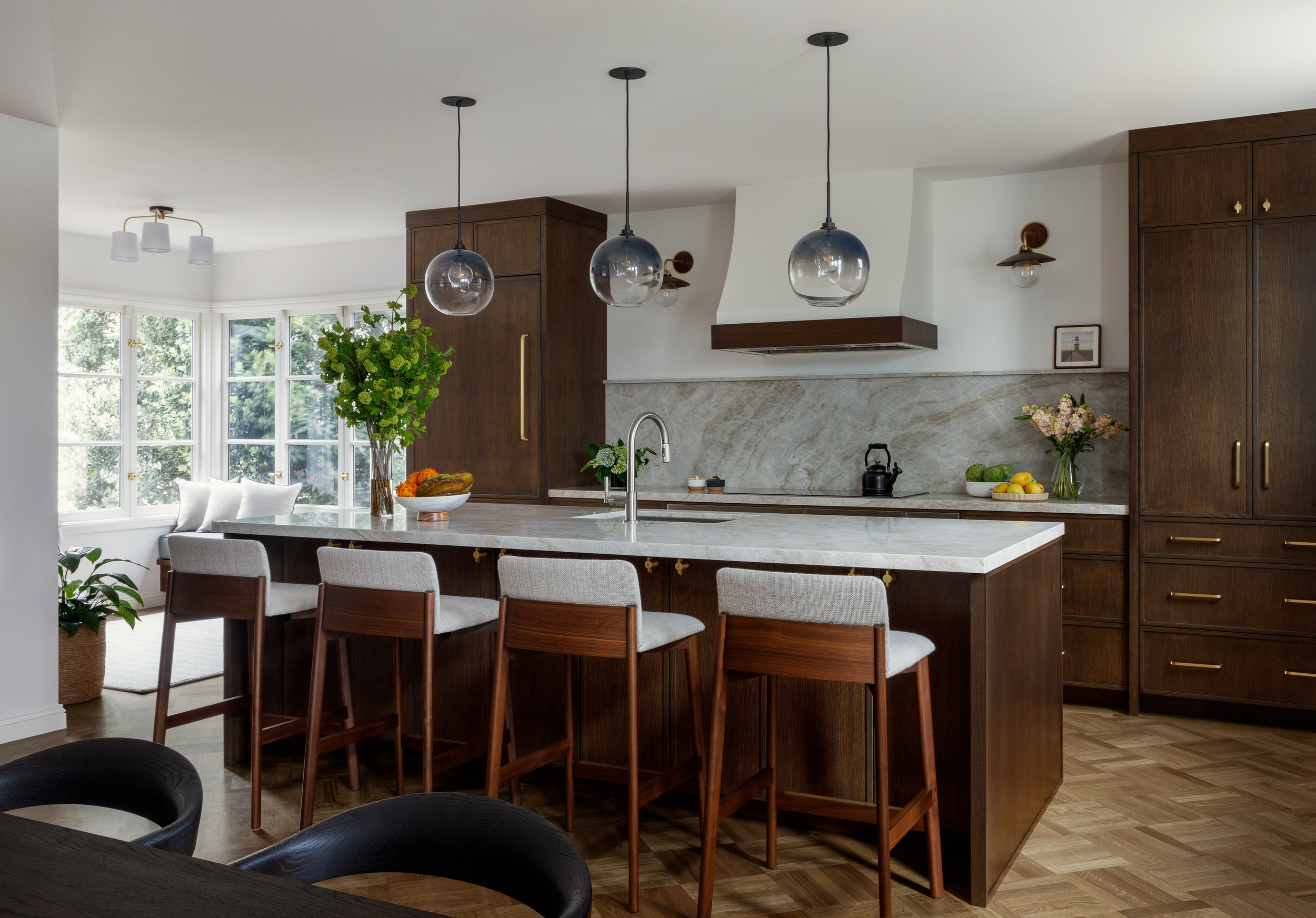 Modern kitchen interior with wooden cabinets, white countertops, and island seating. Features pendant lights, a window nook with cushions, and decorative plants.