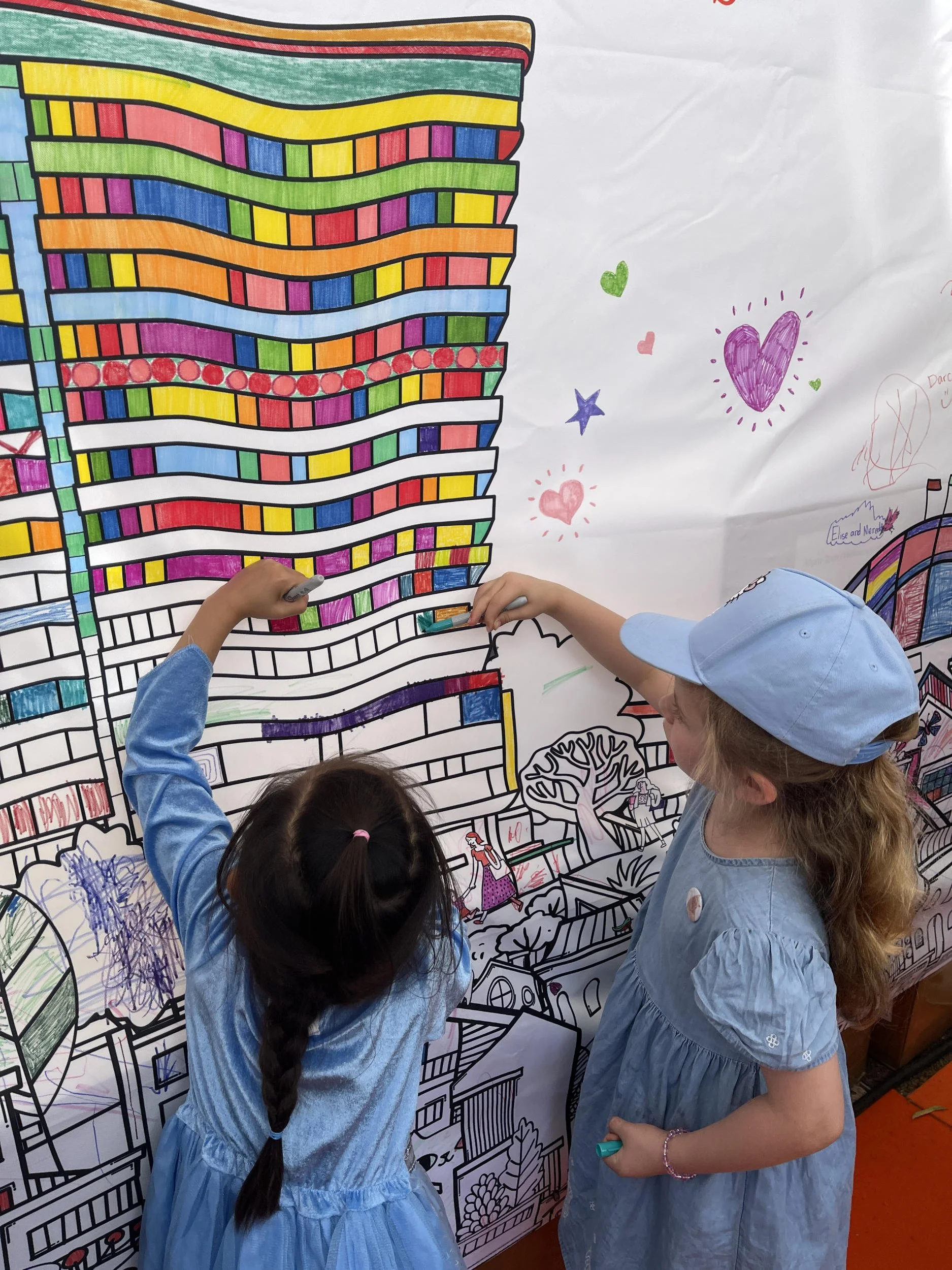 Children coloring a large-scale participatory coloring mural by Aly Indermuhle — a hand-drawn cityscape installation created for a community festival.
