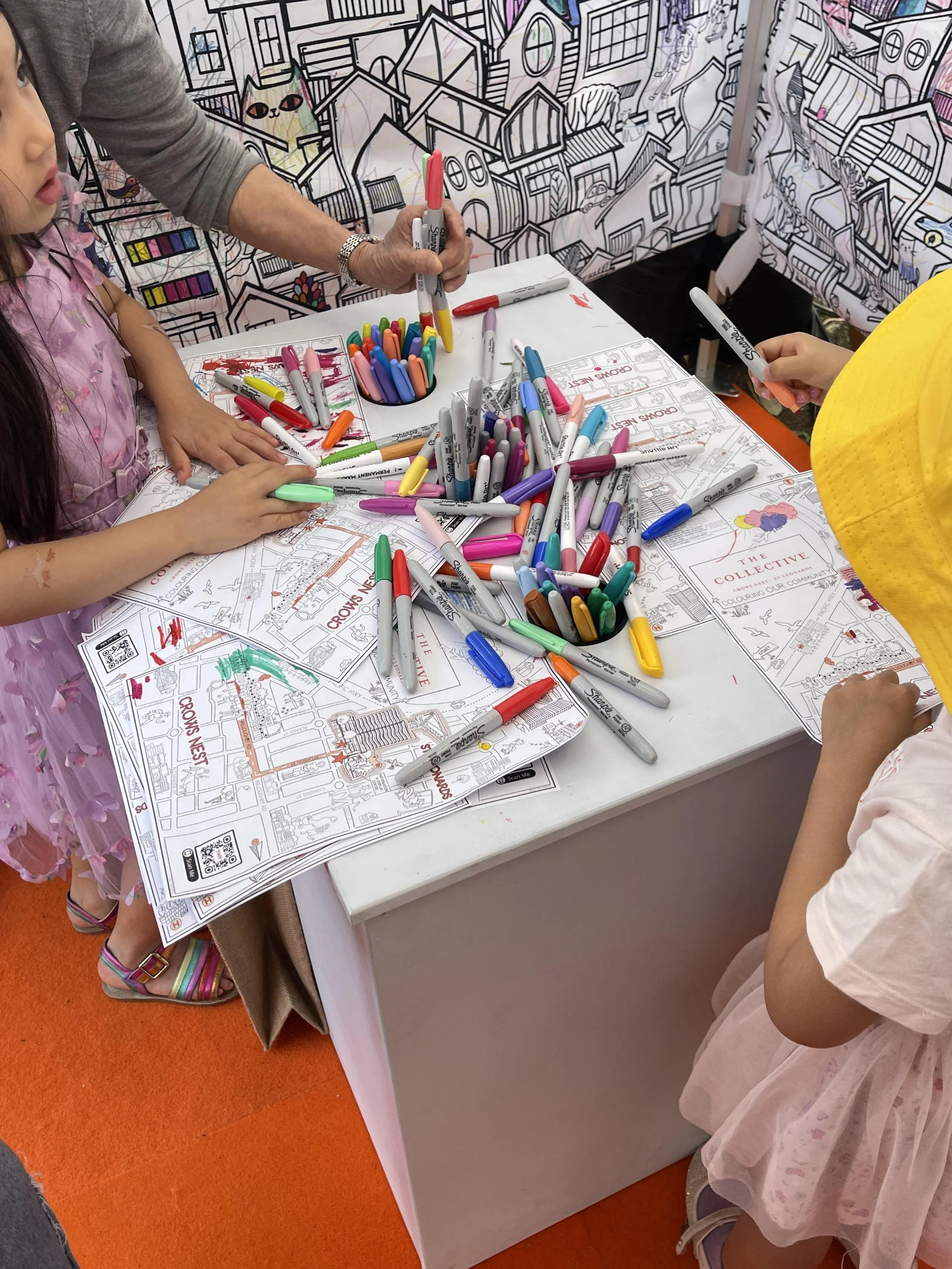 Participants coloring a hand-drawn wayfinding map and interacting with a large-scale participatory coloring mural by Aly Indermuhle at a public community event.
