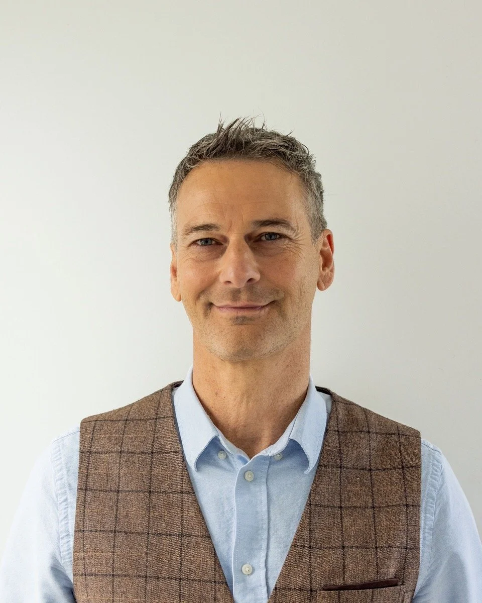 A middle-aged man with short, gray hair smiles slightly and looks at the camera, wearing a light blue shirt and a brown checkered vest against a plain white background.