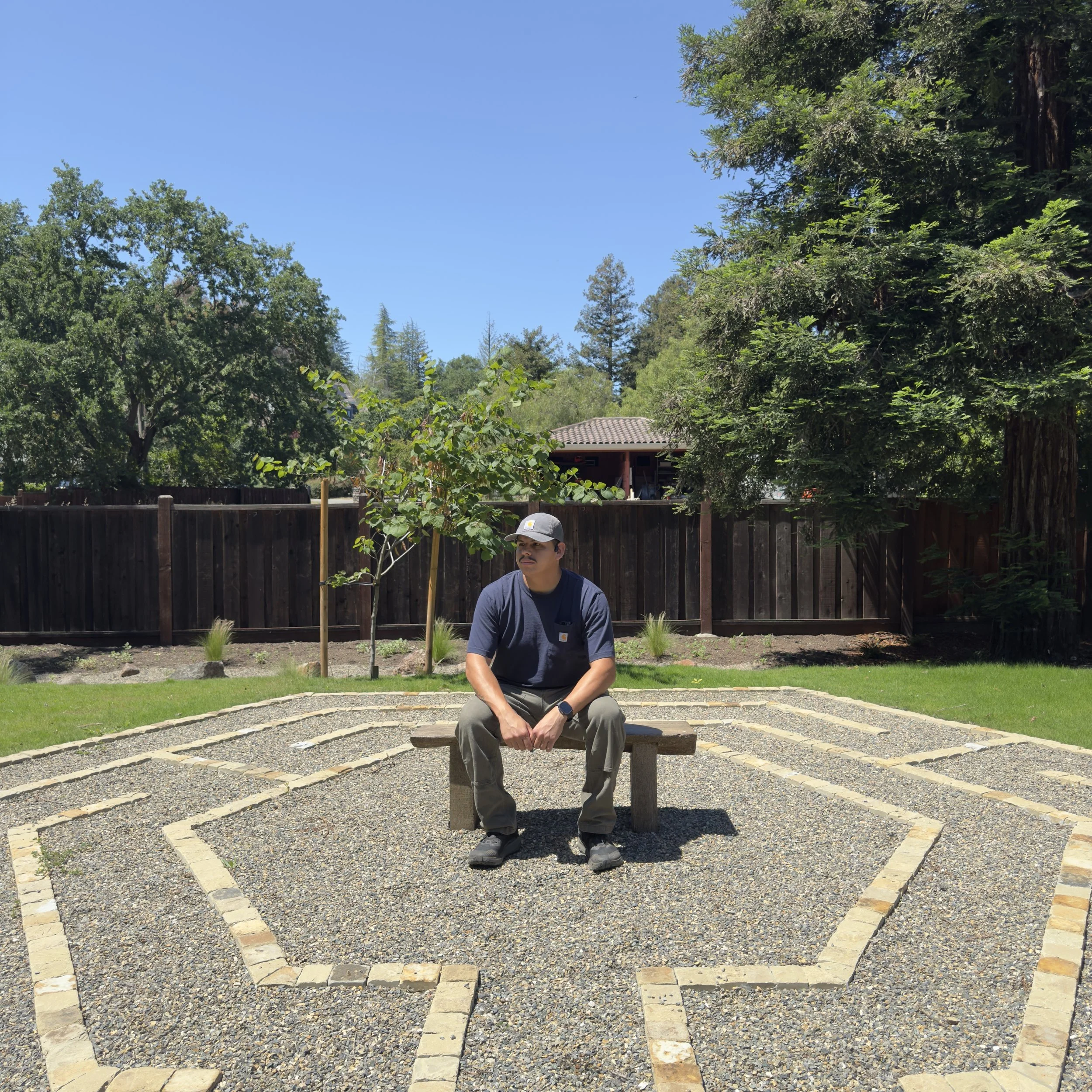 A man in gray clothing with a hat sitting on a wooden bench in a backyard, surrounded by a landscaped garden with newly planted trees, a dark wooden fence, and a blue sky overhead.